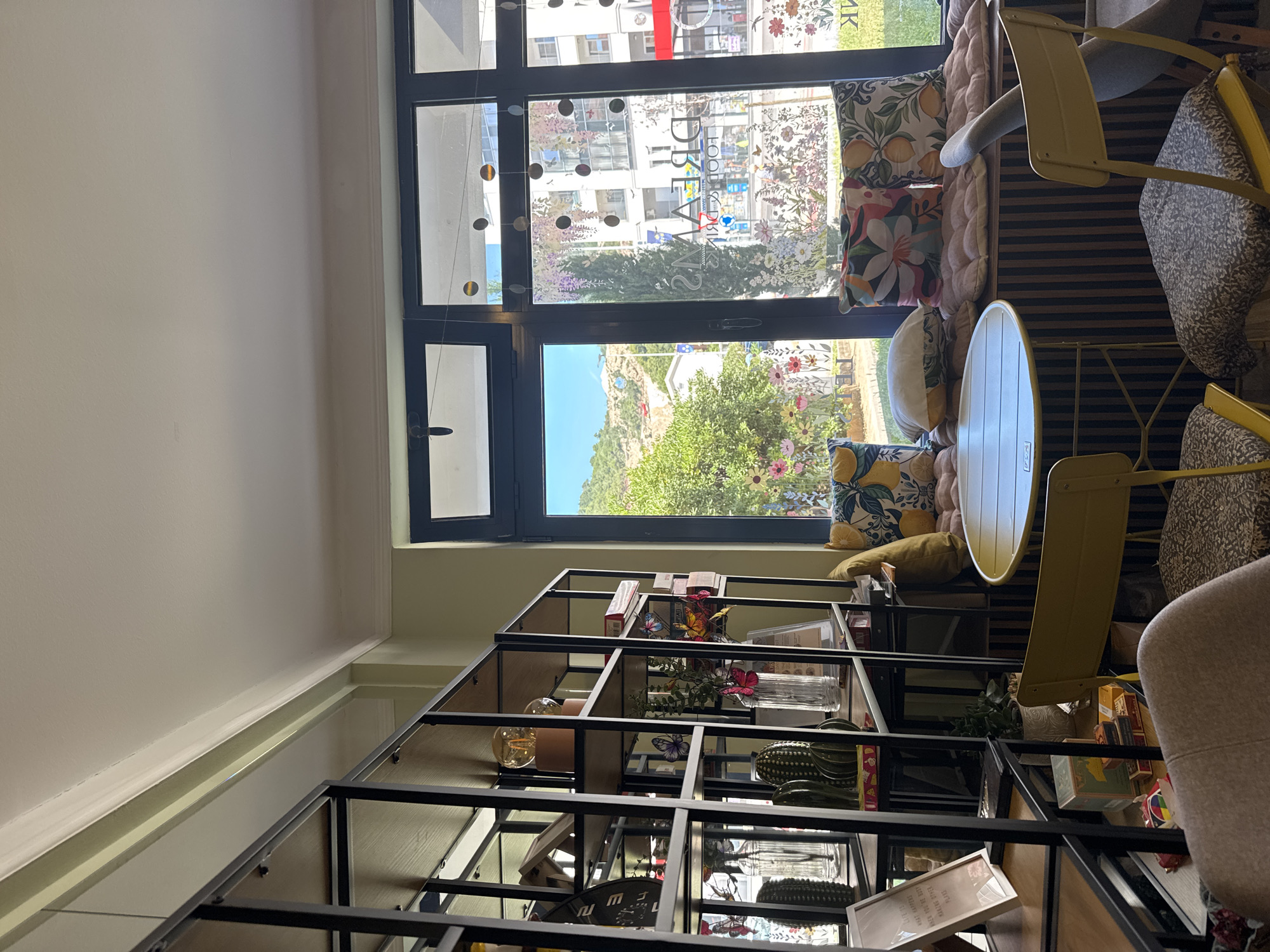 The image displays the interior of "Dreams Food & Drinks," a cafe situated in Budva, Montenegro, viewed from within and looking outward through a series of large, dark-framed windows. The interior features a light-colored ceiling with a white crown molding and light green walls. A black metal shelving unit with multiple wooden shelves dominates the left side of the frame, holding diverse decorative items: prominent exposed-filament light bulbs on wooden bases, artificial green cacti, various clear glass vases containing artificial flowers and colorful butterfly ornaments, framed text, and an assortment of small boxes and decorative objects.
In the mid-ground, a small, round yellow metal table is present, flanked by two yellow metal chairs, each equipped with a patterned fabric seat cushion. A beige upholstered armchair is partially visible in the lower left corner. Beneath the windows, a window seat or cushioned bench is established, featuring vertically striped paneling underneath and adorned with numerous cushions displaying floral patterns (lemons, tropical leaves), plain white covers, and quilted pink/peach designs.
The large windows themselves are segmented into multiple panes and feature decorative strings of circular elements suspended from the top, along with floral and leafy decals applied to the lower glass sections. Through the windows, an exterior scene is visible. The left portion reveals a vibrant green tree in the immediate foreground, a street beyond, and a distant vegetated hill showcasing several light-colored structures and a distinct reddish architectural element. The right portion of the exterior view displays a more urban setting with multi-story buildings, numerous windows, and a tall, slender tree. The reversed text "DREAMS FOOD & DRINKS" is clearly legible on one of the central windowpanes. No individuals are visible in the scene.