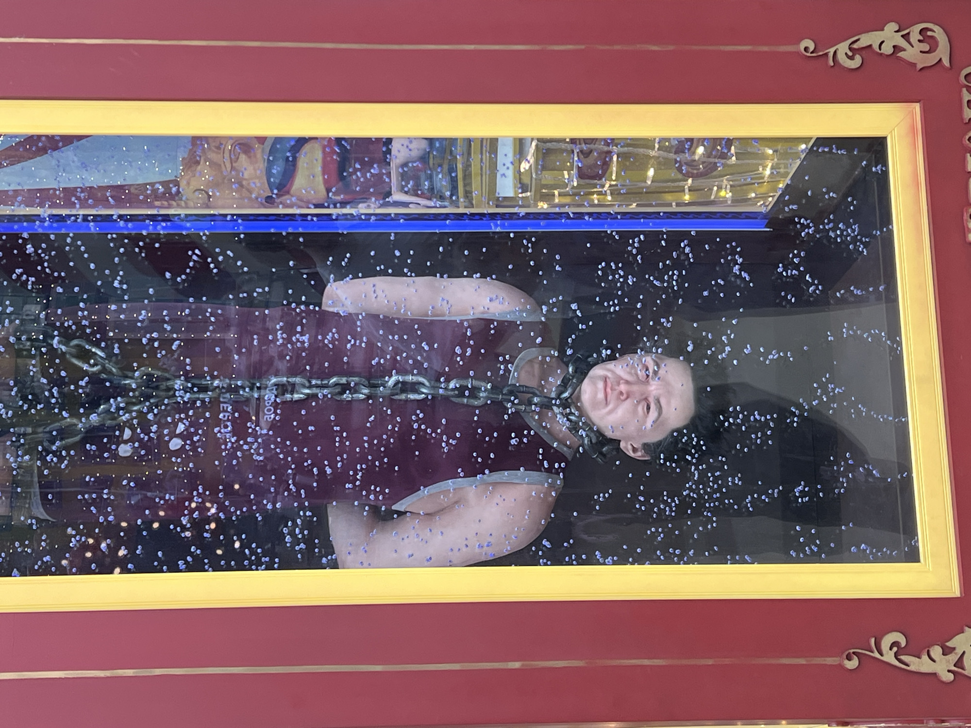 A person is suspended upside down by a chain inside a glass enclosure, submerged in water with numerous bubbles. The person is wearing a dark garment. The enclosure is framed by red and gold trim, with decorative elements on the red exterior. In the background, behind the person, there is a colorful, possibly circus-themed, backdrop and a structure with lights. The scene appears to be an attraction or exhibit. The location is the Moscow Circus on Tsvetnoy Boulevard, Moscow, Russia.