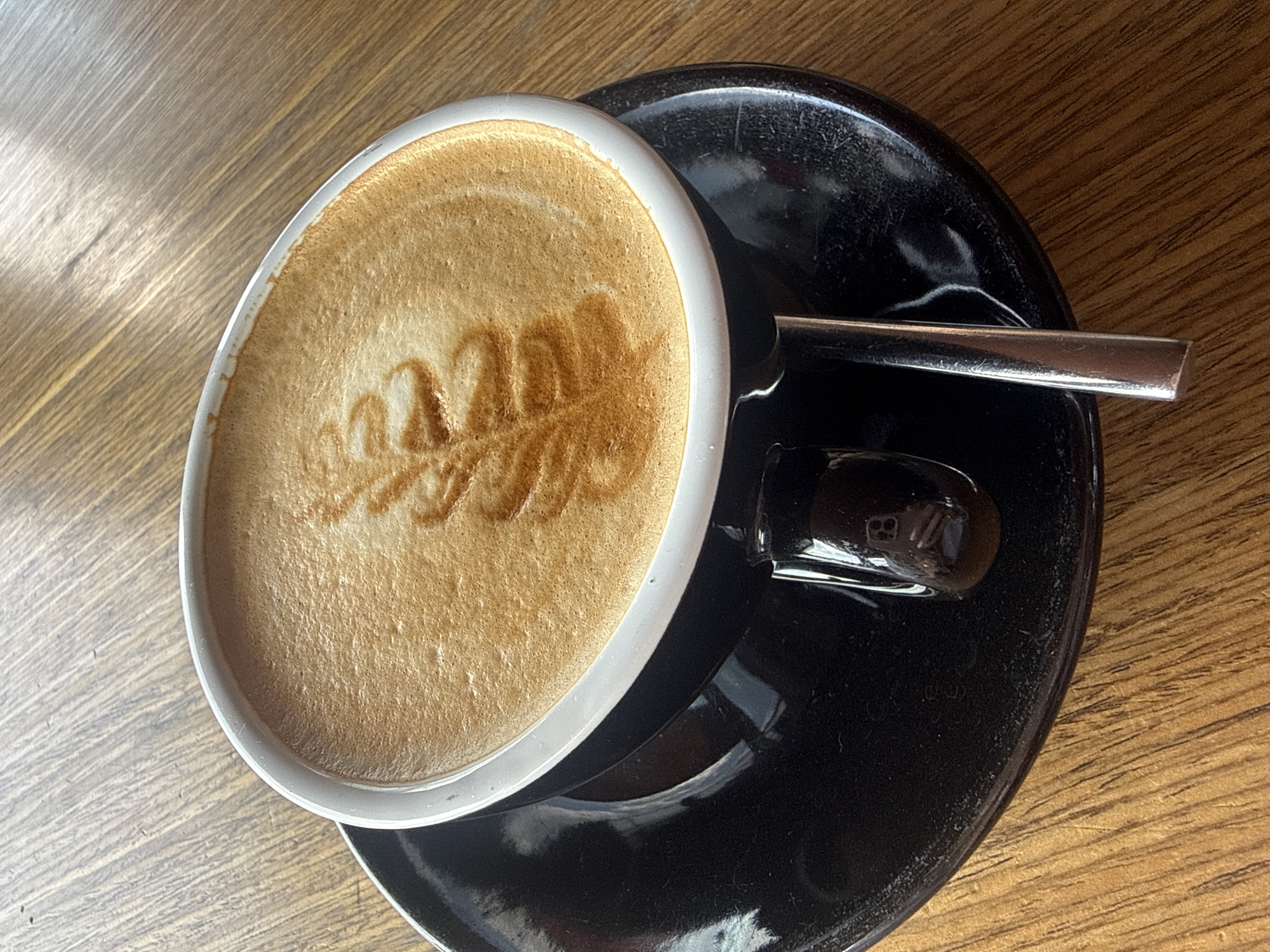 A close-up, overhead shot shows a white ceramic mug of coffee with latte art, placed on a black saucer. The coffee has a foamy, light brown surface with a darker brown pattern resembling a leaf or feather. A silver spoon rests on the saucer, partially submerged in the coffee. The mug and saucer sit on a wooden surface with visible grain. The scene appears to be indoors, with natural light illuminating the coffee and creating reflections. There are no people or animals visible in the image. This image was taken in the city of Kerch, Ukraine.