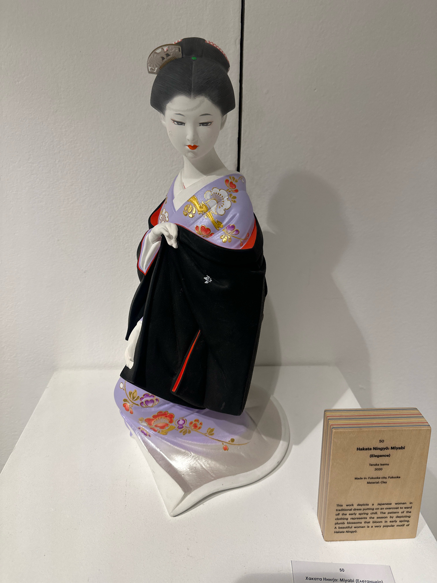 The image shows a traditional Japanese doll displayed on a white pedestal in what appears to be an exhibition space. The doll, identified as "Hakata Ningyō: Miyabi (Elegance)" by Tanaka Isamu, is dressed in a purple and white kimono with gold floral patterns and a black outer garment. Beside the doll, a small wooden plaque with text in English and what appears to be Cyrillic describes the artwork and its origins in Fukuoka, Japan. The background is a plain white wall, with shadows cast on it from the display. No people or specific city landmarks of Belgrade, Serbia are visible in this image.