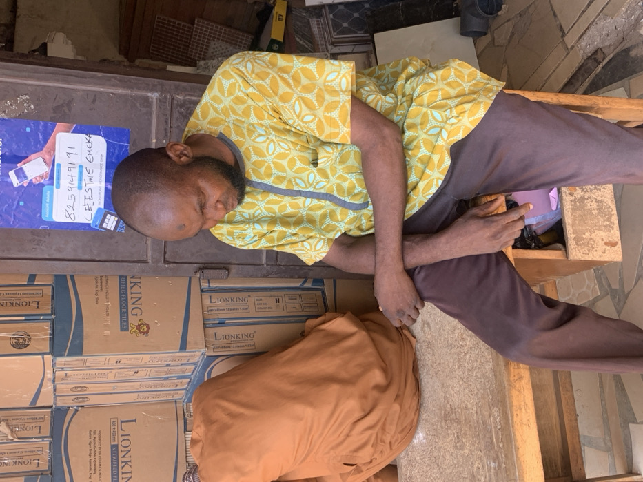 In a retail or storage area within Magongo, Nigeria, a single male individual is seated on a wooden bench. The man, with dark skin, a short beard, and a bald head displaying a small dark mark on the forehead, wears a short-sleeved yellow patterned shirt with a grey vertical stripe and dark brown trousers. He is positioned facing slightly right, looking downwards, with his arms crossed. To his immediate left on the same bench, a large object covered in brown fabric is visible. Behind the man, to the left, is a stack of cardboard boxes labeled "LIONKING VERIFIED FLOOR TILES" with "MADE IN NIGERIA" text. Directly behind him, on a dark metallic door or shutter, a blue poster is affixed, displaying "Account details," a phone number "8259149191," the name "CELESTINE EMEKA," and "ACCESS BANK." Beneath the bench, a small wooden compartment holds a pink plastic bag and other items. The foreground shows a light-colored tiled floor, and some loose tiles are visible on the right. The scene is illuminated by ambient daylight.
