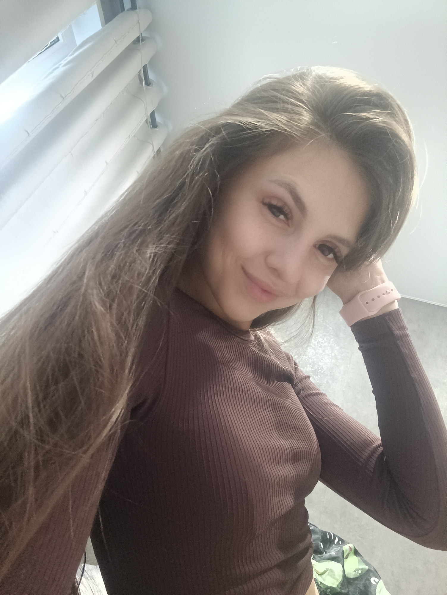 A young woman with long, brown hair is pictured in a selfie. She is wearing a brown, ribbed long-sleeved shirt and a pink smartwatch. Her right arm is bent, with her hand resting near her head. She is smiling gently. The background includes white blinds and a wall. The image appears to be taken indoors.