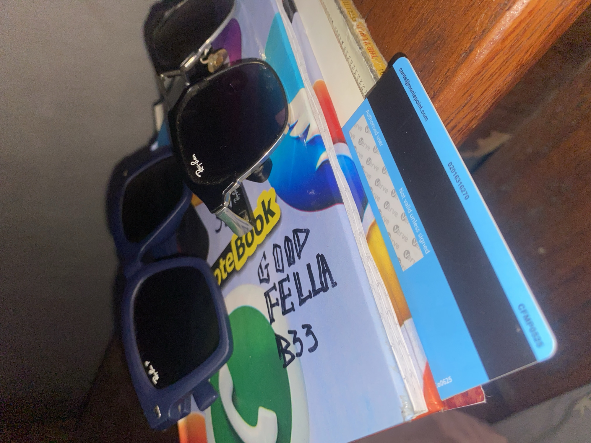 The image captures a collection of personal items arranged on a wooden surface, possibly a shelf or desk, in an indoor setting. The shot is taken at a tilted angle, looking down at the objects.
In the foreground, there are two pairs of sunglasses. The top pair is black with "Ray-Ban" visible on the right lens, while the bottom pair is blue, also featuring a "Ray-Ban" logo on its right lens. Behind the sunglasses is a light-colored notebook with "NoteBook" printed on its cover and a large green WhatsApp logo. Handwritten in black ink on the notebook are the words "GOOD FELLA B33".
To the right of the notebook, leaning against other unseen books or papers, is a blue and black financial card. Visible text on the card includes an email address "cards@moniepoint.com", a number "02016316270", "Authorised user", "Not valid unless signed", and multiple instances of the "Verve" logo. Another code, "CFMP0525", is partially visible on the bottom right edge of the card.
No activity is depicted, and no people are present. The lighting suggests an indoor environment. Given the presence of "moniepoint.com" and "Verve" branding, which are associated with Nigerian financial services, the items strongly indicate a location context in Nigeria, specifically Jama A as provided.