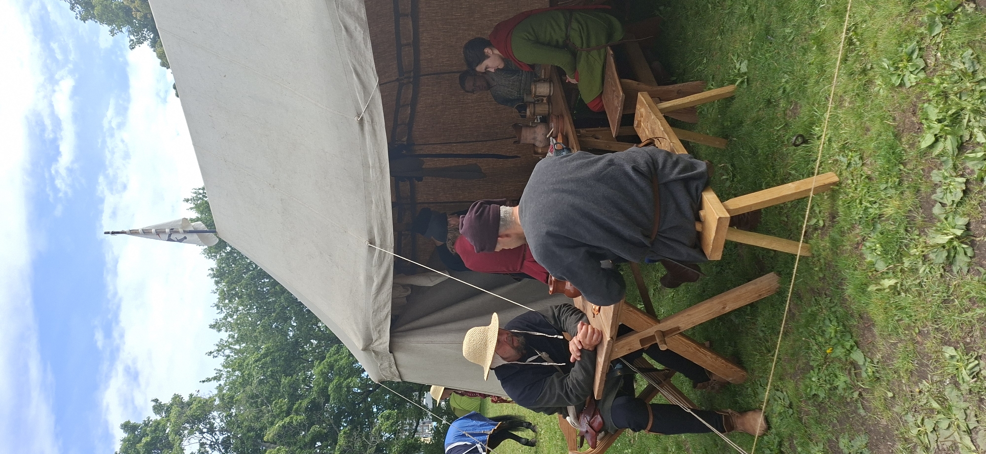In the city of Turku, Finland, on a grassy outdoor area, a reenactment event is taking place. In the foreground, two men in historical attire are seated at a wooden picnic table. The man on the left wears a straw hat, a dark tunic, and leather boots, with a leather pouch and sword at his hip. The man on the right wears a dark cap and a dark grey tunic. They are facing each other, with their hands resting on the table, suggesting a paused conversation or the beginning of a game.
Behind them, a large canvas tent provides shade. Inside the tent, several other individuals are visible, also dressed in historical clothing. A woman in a green tunic and red headscarf sits on a bench, facing away from the camera, interacting with another person. A man in a red tunic stands near the entrance of the tent. Further back, more people are obscured by the tent's structure. A white flag with a dark symbol is attached to a pole at the peak of the tent.
To the left of the tent, a horse with a blue blanket is visible, suggesting the presence of cavalry or transportation relevant to the historical period being reenacted. The environment is grassy, with trees and a cloudy sky forming the backdrop. The scene depicts a gathering or encampment with a focus on historical interpretation.