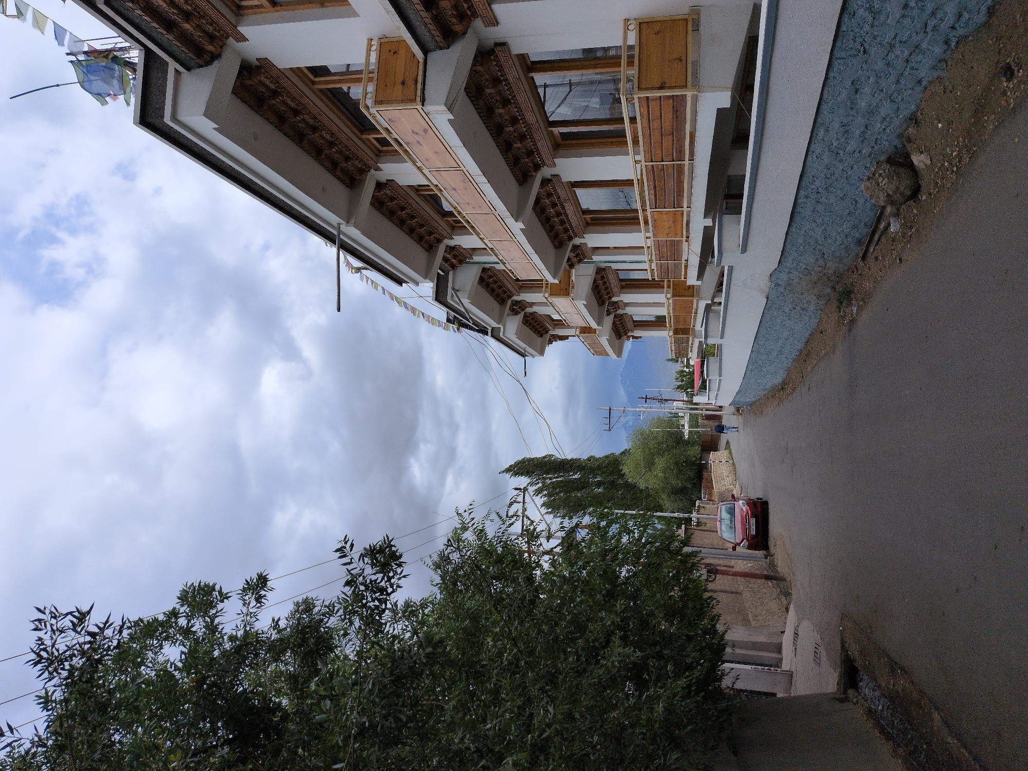 A red car is parked on the side of a paved road in Leh, India. The road is lined with buildings and trees. A person is walking down the road. The sky is cloudy.