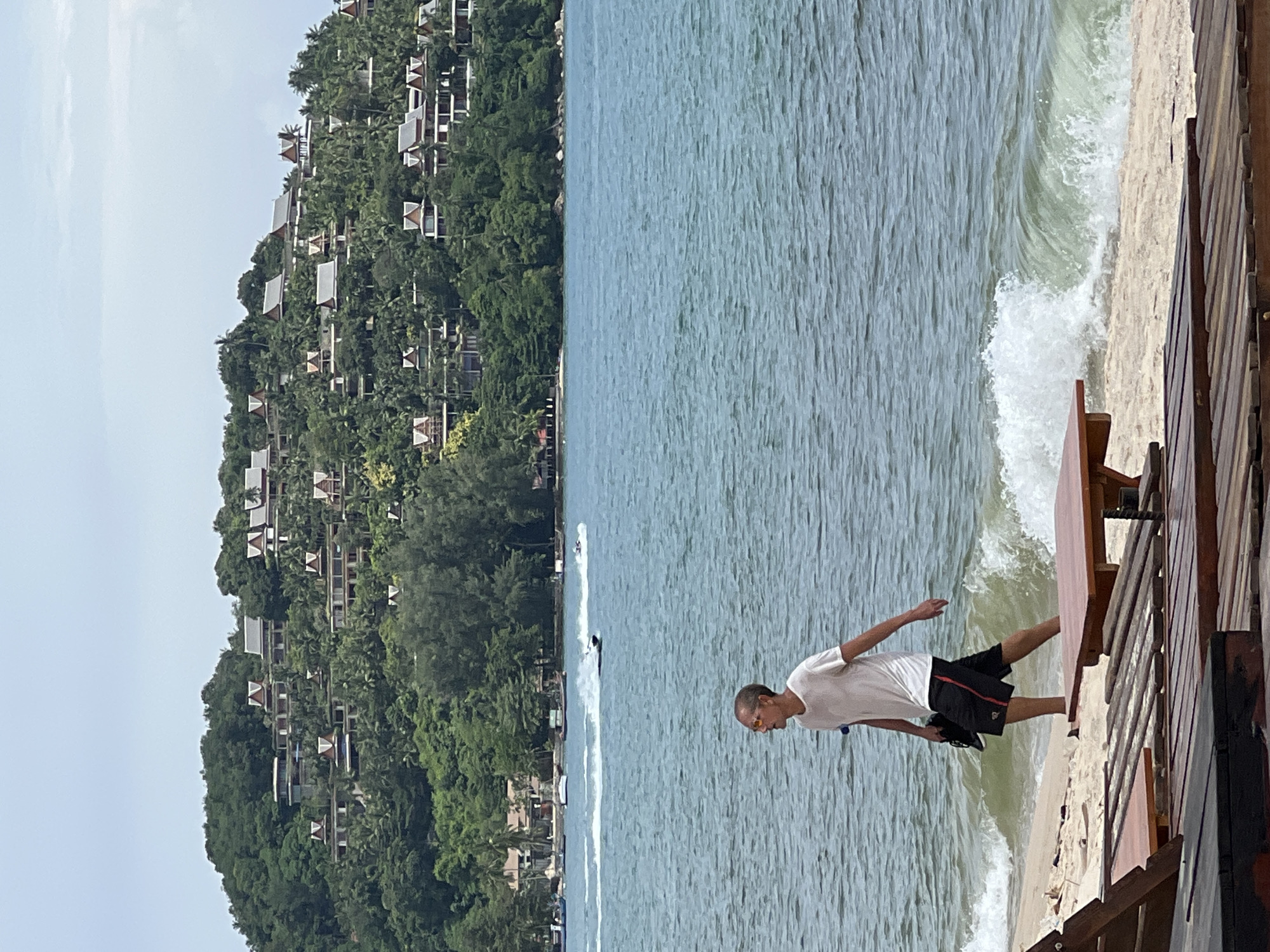 A man wearing a white t-shirt and black shorts with red stripes walks along a sandy beach towards the right side of the frame. He is holding a pair of dark shoes in his left hand. Gentle waves break on the shore around his ankles. In the foreground, wooden lounge chairs and a small table are partially visible. In the background, the blue ocean stretches to the horizon, with several boats and jet skis visible in the distance. Across the water, a lush green hillside is covered with numerous villas and tropical trees, topped with a hazy sky.