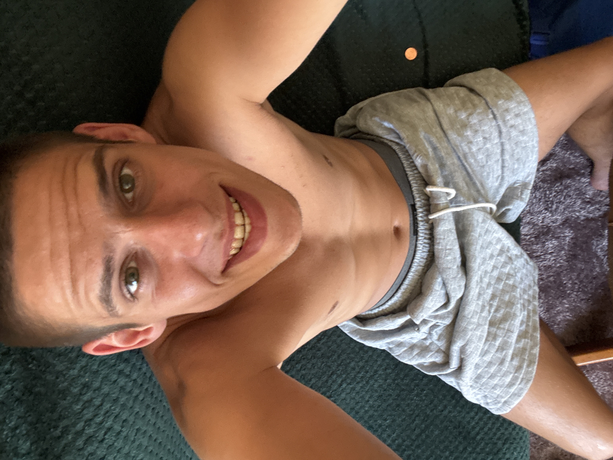 A man with short brown hair and green eyes smiles at the camera, looking down from a high angle. He is shirtless, wearing gray athletic shorts. His skin is tan, and he has a visible mole on his chest. The background is a dark green textured surface, possibly a couch or carpet, with a small copper-colored object, possibly a coin, visible to the right of the man's torso. The man's legs and knees are bent. The lighting is natural and comes from above.