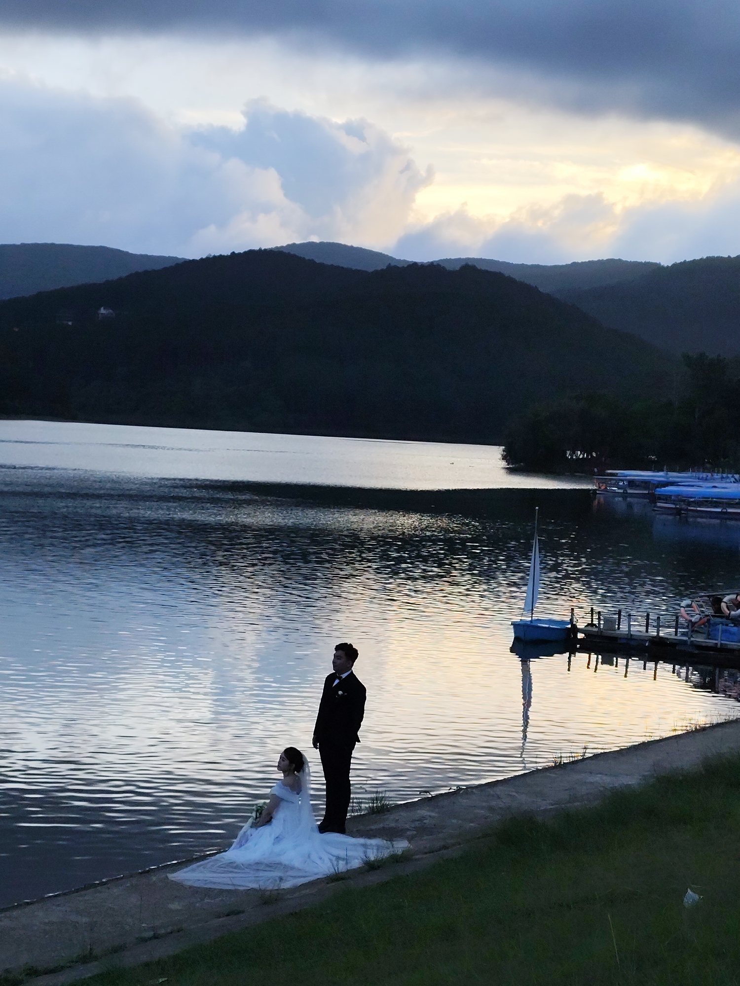 A full-length shot shows a couple posing by a lake. The woman is sitting on the grassy bank wearing a white wedding dress. The man is standing next to her, wearing a dark suit and tie. Mountains are visible in the background. The sky is overcast. Reflections can be seen on the lake water. A small boat sits next to a dock on the lake. The location is in Da Lat, Vietnam.
