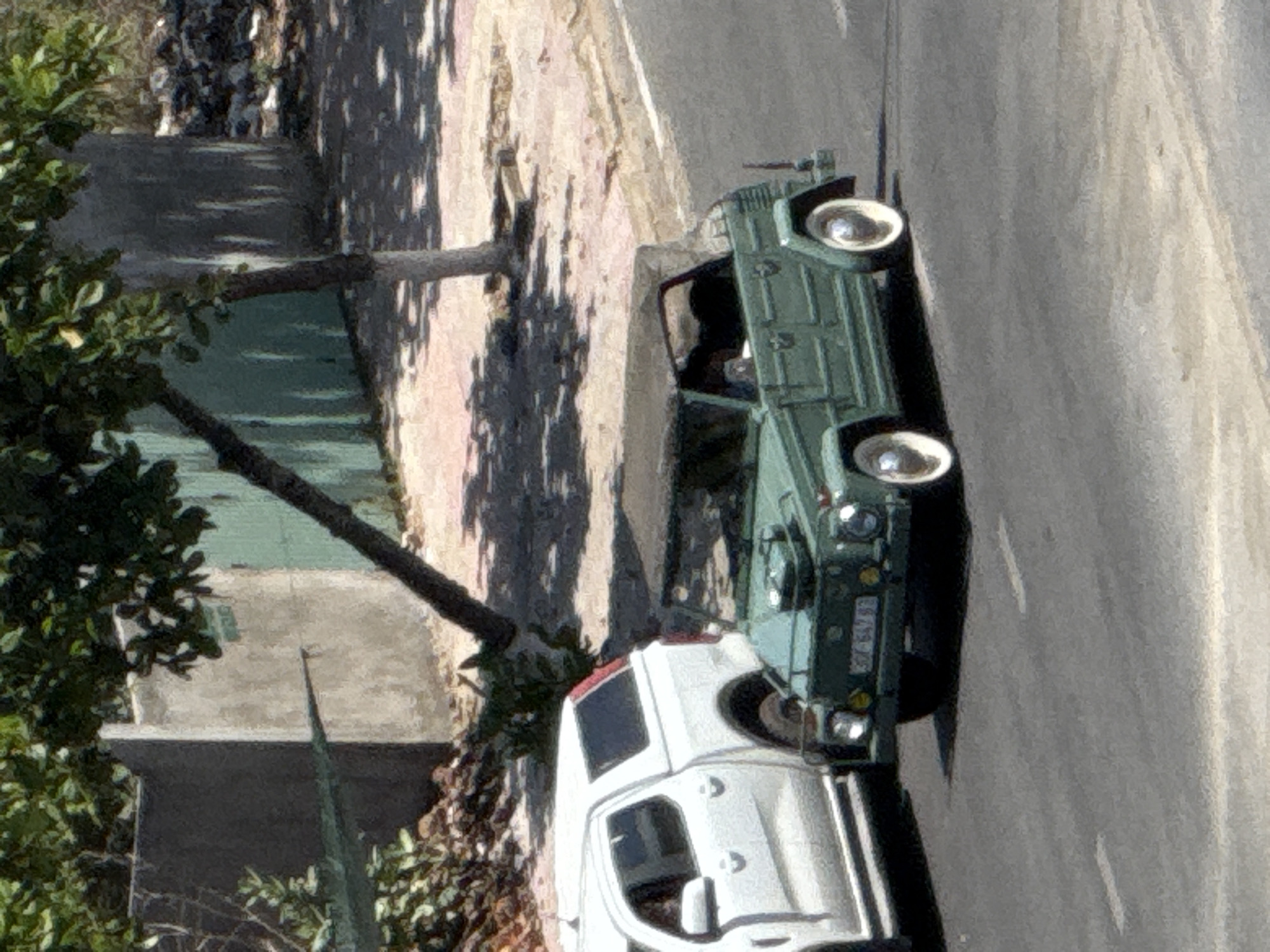 Two vehicles are parked on a paved road, one behind the other. The front vehicle is a vintage green car with a light-colored roof. It has round headlights and a license plate that appears to read "37E 5015". The second vehicle, parked directly behind the green car, is a white van. The scene is illuminated by bright sunlight, casting distinct shadows on the ground and road, suggesting it is daytime. The surrounding environment includes lush green foliage on the left and a light-colored building or wall on the right. The image is captured from a slightly elevated perspective. No people are visible in the frame. The overall setting suggests a warm, sunny day in a location with a mix of urban and natural elements.