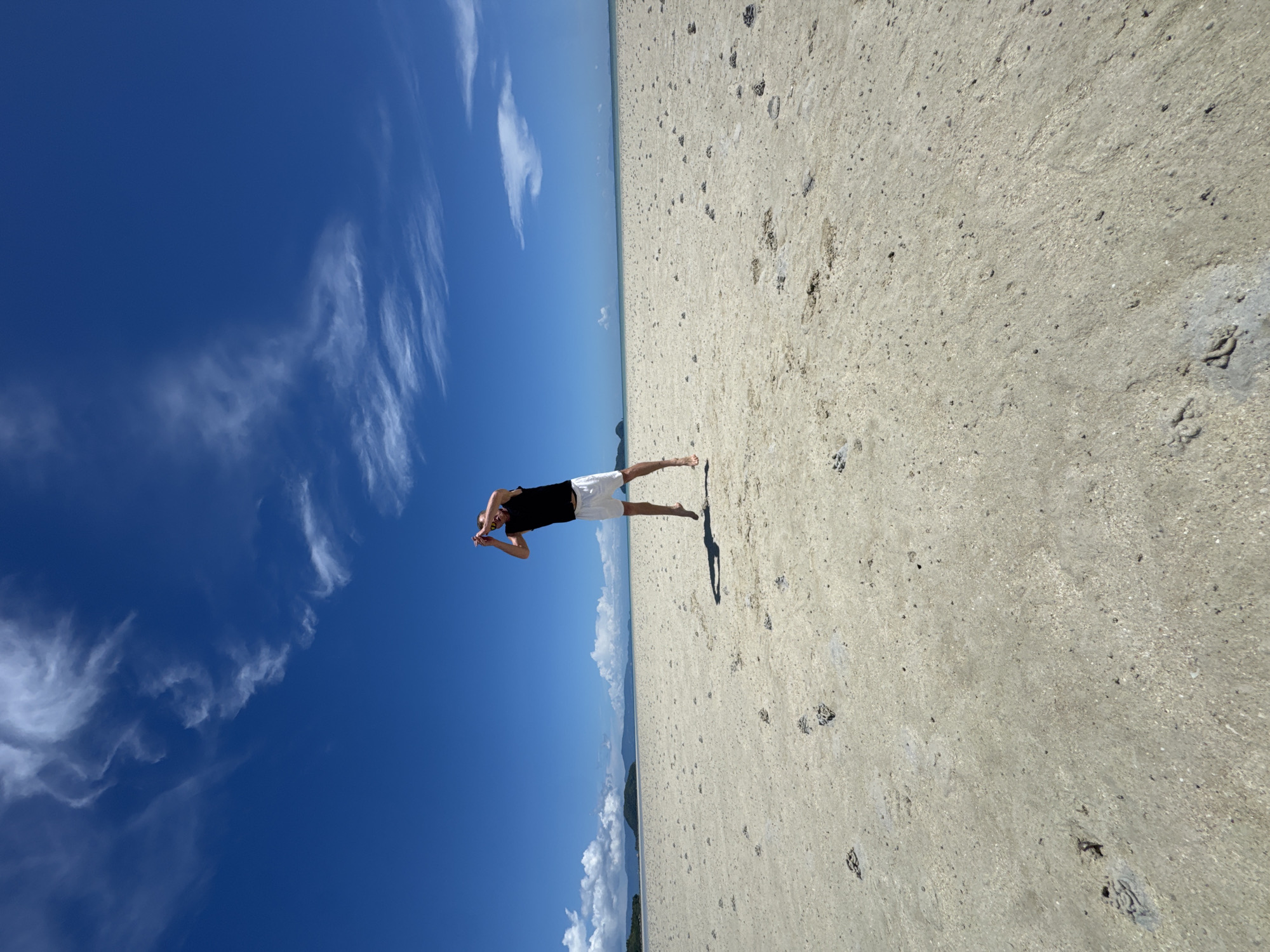 A person is captured mid-air, jumping on a vast, sandy beach under a bright blue sky with wispy clouds. The person is wearing a black t-shirt and white shorts. The beach appears to be low tide, revealing numerous small, dark mounds scattered across the sand, indicative of crab holes. In the distance, a thin strip of land with vegetation, possibly islands, is visible along the horizon, separating the sandy expanse from the sea and sky. The overall scene suggests a remote and tranquil coastal environment on Koh Phangan, Thailand.