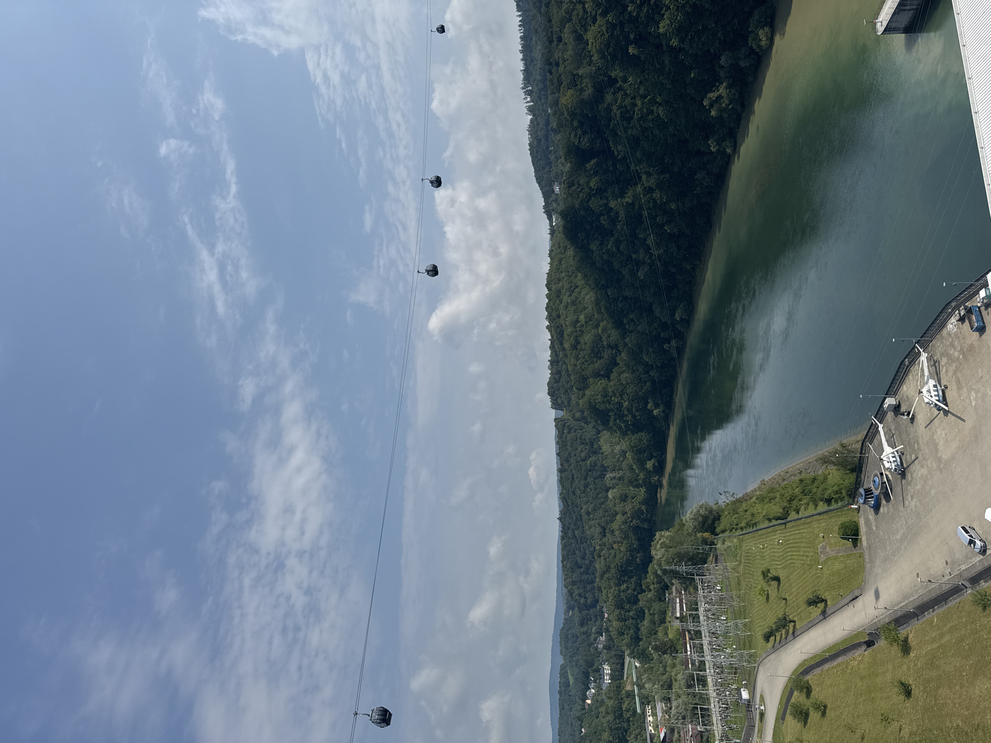 The image shows a wide river flowing through a lush, forested landscape. The sky is blue with scattered white clouds. Several gondolas are suspended on wires, crossing the frame from left to right. On the left bank of the river, there is a power station with numerous tall transmission towers. Adjacent to the power station, a paved area is visible with several parked vehicles, including two helicopters and a van. The overall scene suggests a hydroelectric dam or similar industrial facility integrated into a natural environment, possibly near Solina, Poland, given the presence of a dam and reservoir often associated with that location.