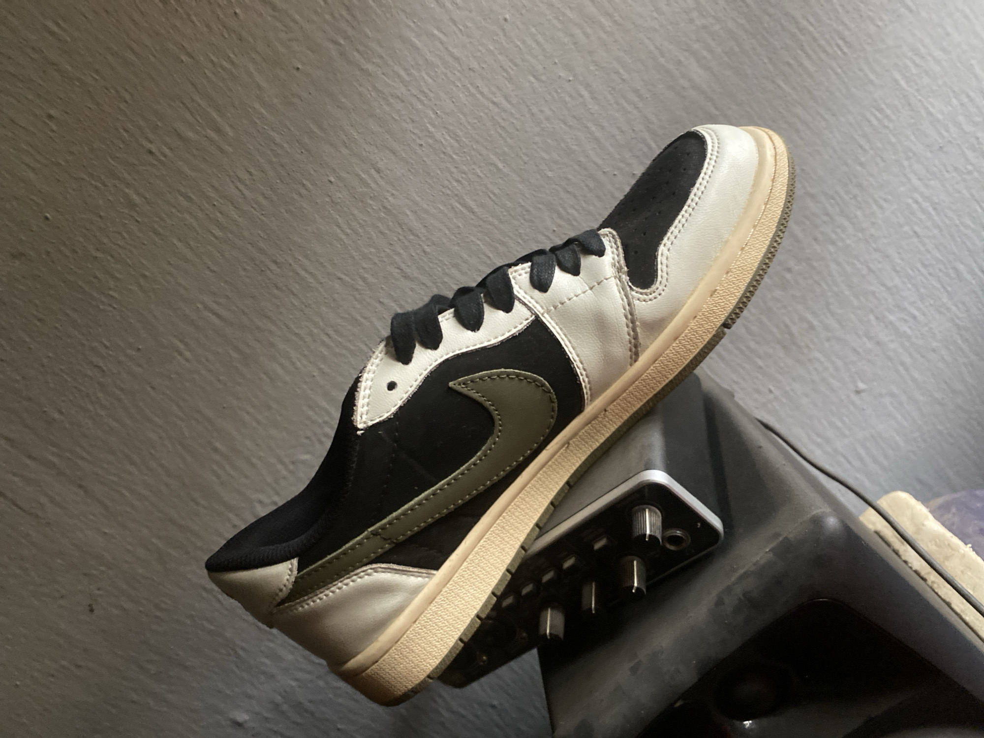 A single black and white athletic shoe with a tan sole and black laces is balanced on top of a dark gray electronic device with silver knobs. The background is a mottled gray wall. The scene appears to be indoors. The location in the city of Hanwa, Nigeria is not discernible from the image.