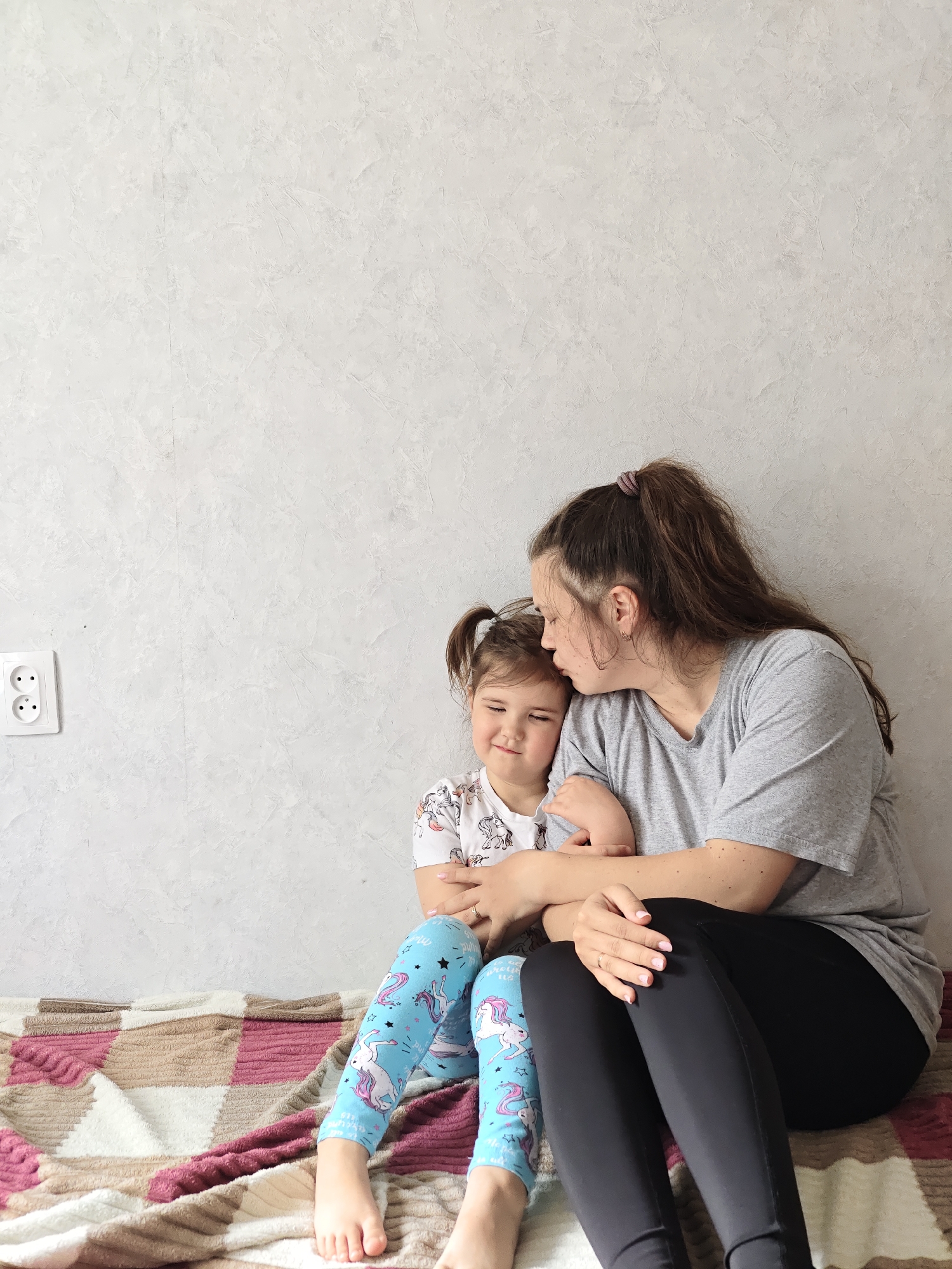 A woman and a young girl are seated on a patterned blanket or bedspread against a light gray wall. An electrical outlet is visible on the wall to the left. The woman, who has long, brown hair pulled back in a ponytail, is wearing a gray t-shirt and black leggings. She is embracing the girl and appears to be kissing her forehead. The young girl is wearing a white t-shirt with a unicorn design and blue leggings with a similar unicorn pattern. The location is inferred to be Chaikovsky, Russia.