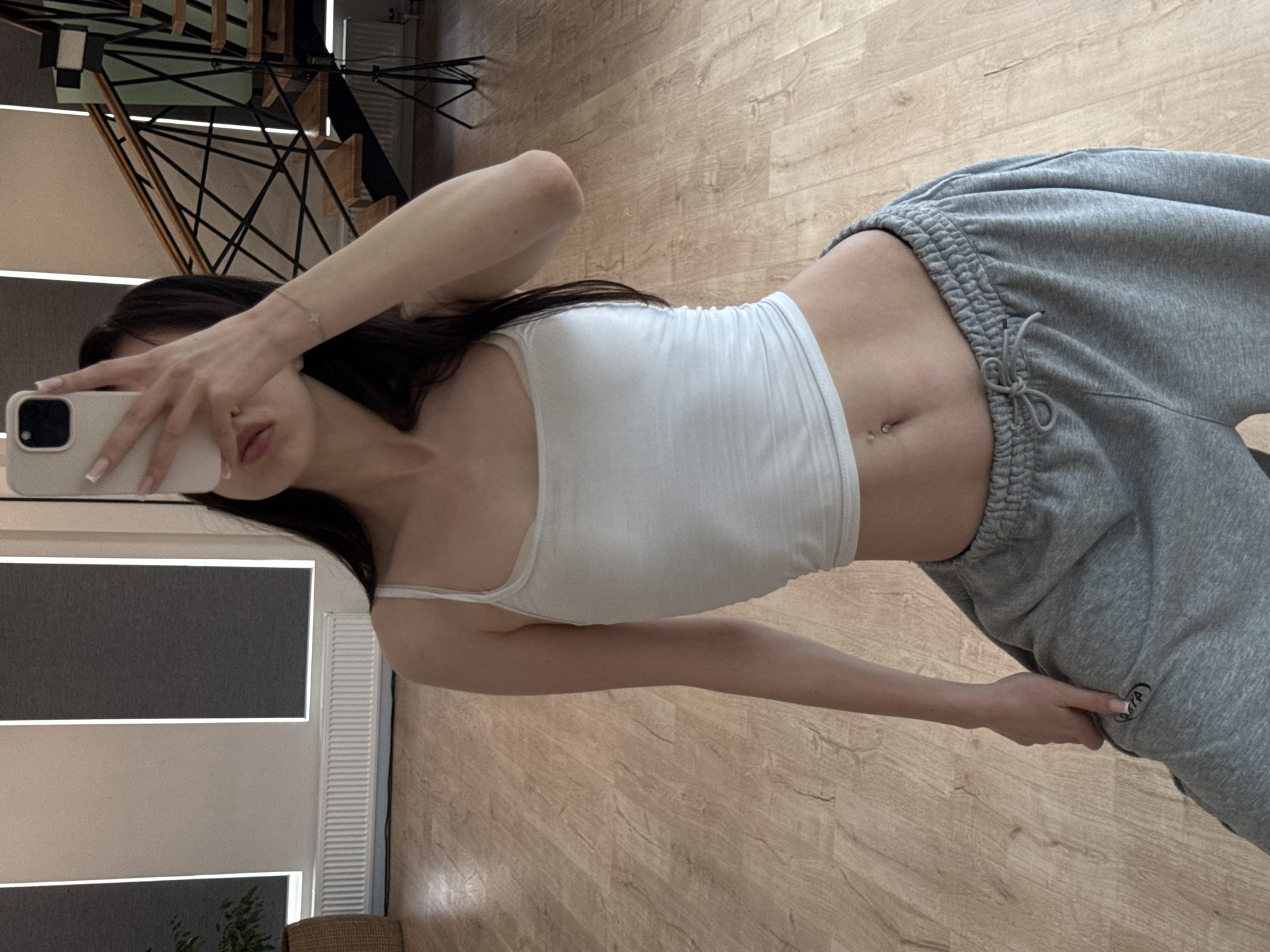 A young woman is taking a selfie in a room with wooden floors. She is wearing a white crop top and grey sweatpants. Her hair is dark and falls over her shoulders. She has a bracelet on her left wrist. In the background, there are modern stairs and a window with blinds. The lighting suggests it is daytime. The woman's facial expression is not fully visible as she is holding a phone to take the photo.
