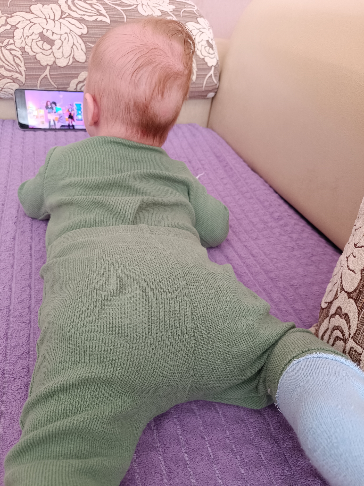 A baby is lying prone on a purple couch, with their head facing a smartphone displaying an image of two animated characters. The baby is wearing a green, ribbed onesie and a blue sock on one leg. The head of the couch has a floral pattern pillow on it. The location cannot be confirmed as Tobolsk, Russia.