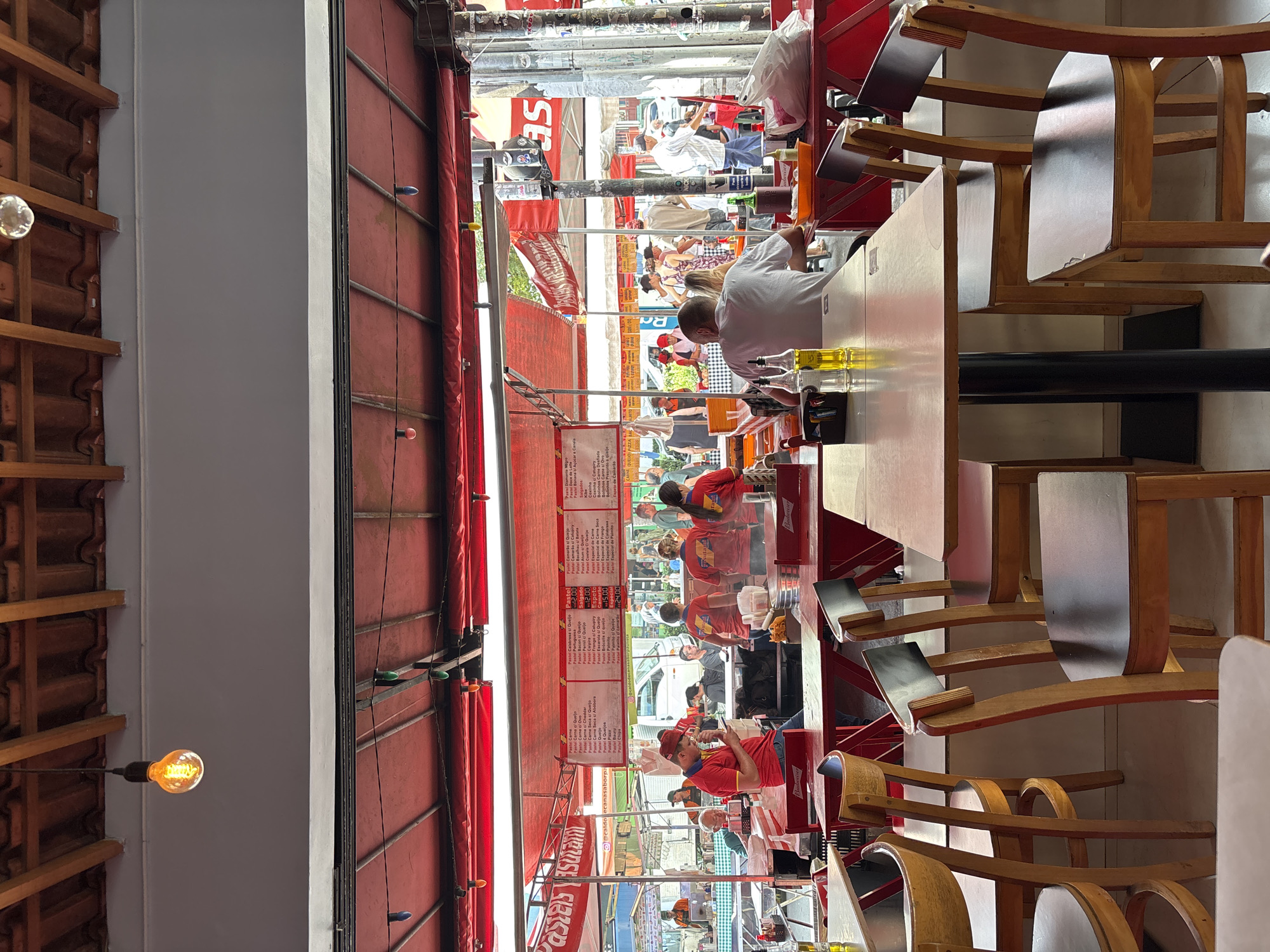 The image shows a street food market in São Paulo, Brazil, specializing in "pastel," a fried pastry. Several individuals are seen eating at a counter under a red awning, and others are visible in the background street scene. Menus displaying various pastel fillings and prices are prominently displayed. There are wooden chairs, tabletops with bottles, and views of the outside street, with people and vehicles. The interior has exposed light bulbs and a textured ceiling.