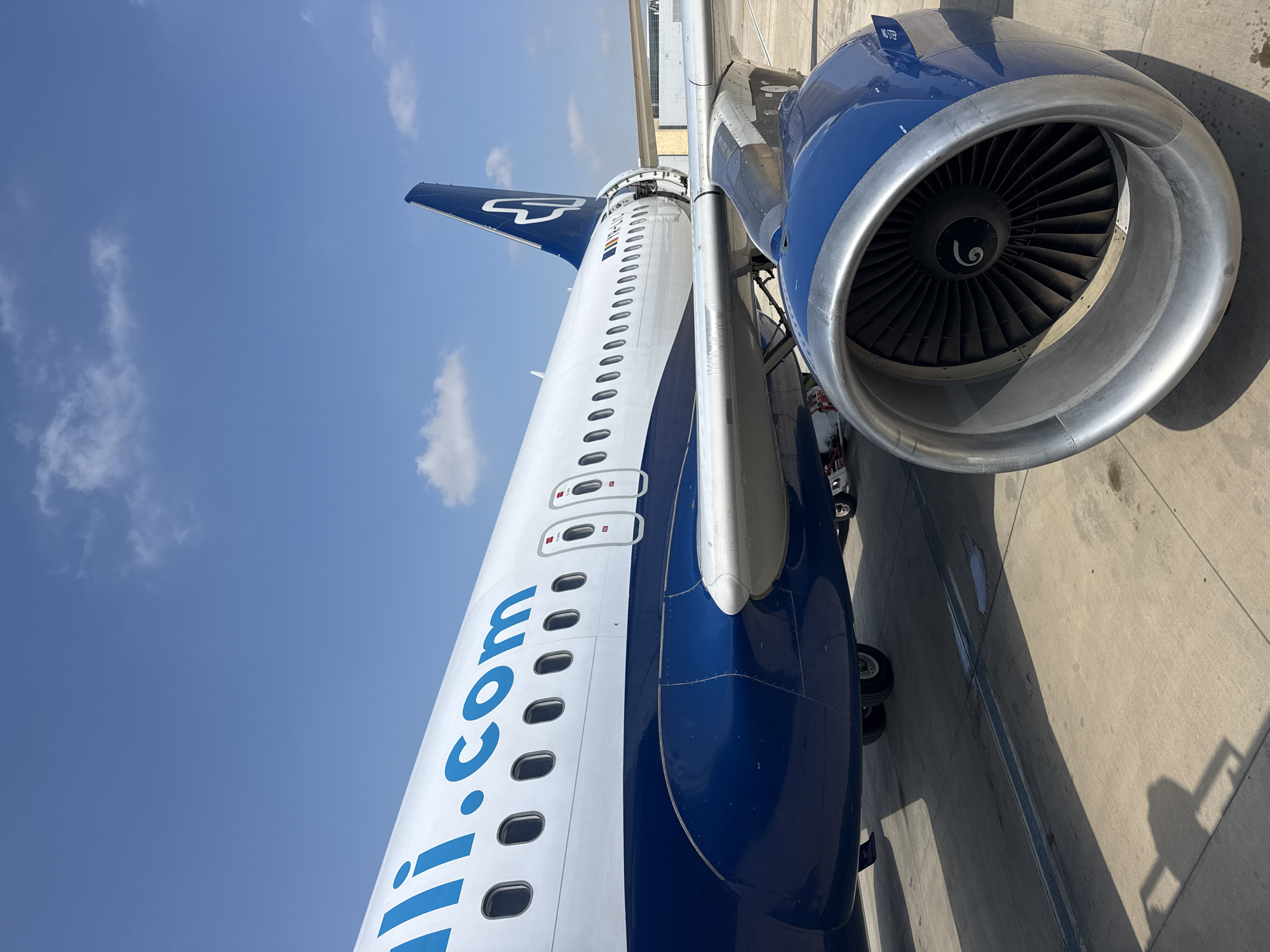 The image shows an aircraft, likely at Sdot Dan Regional Council, Israel. The blue and white fuselage is prominently featured, with the website address "lill.com" visible along the side. An engine is visible on the right. The tail of the aircraft, also blue and white, is visible in the background. The aircraft is parked on a concrete surface. The sky is blue with scattered clouds.