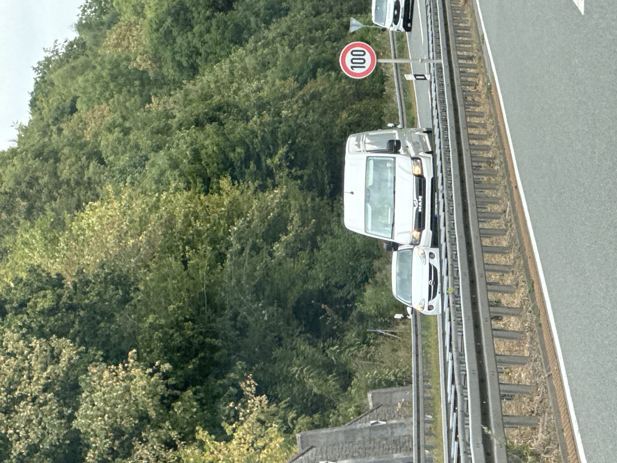 The image shows a view of a highway in Bad Lauterberg im Harz, Germany, with lush green trees lining the side of the road. A white van, labeled "MAN," is driving on the highway, followed by a white car. A speed limit sign indicating "100" is visible to the right of the road. The highway has multiple lanes separated by metal barriers. The scene appears to be a sunny day with good visibility.