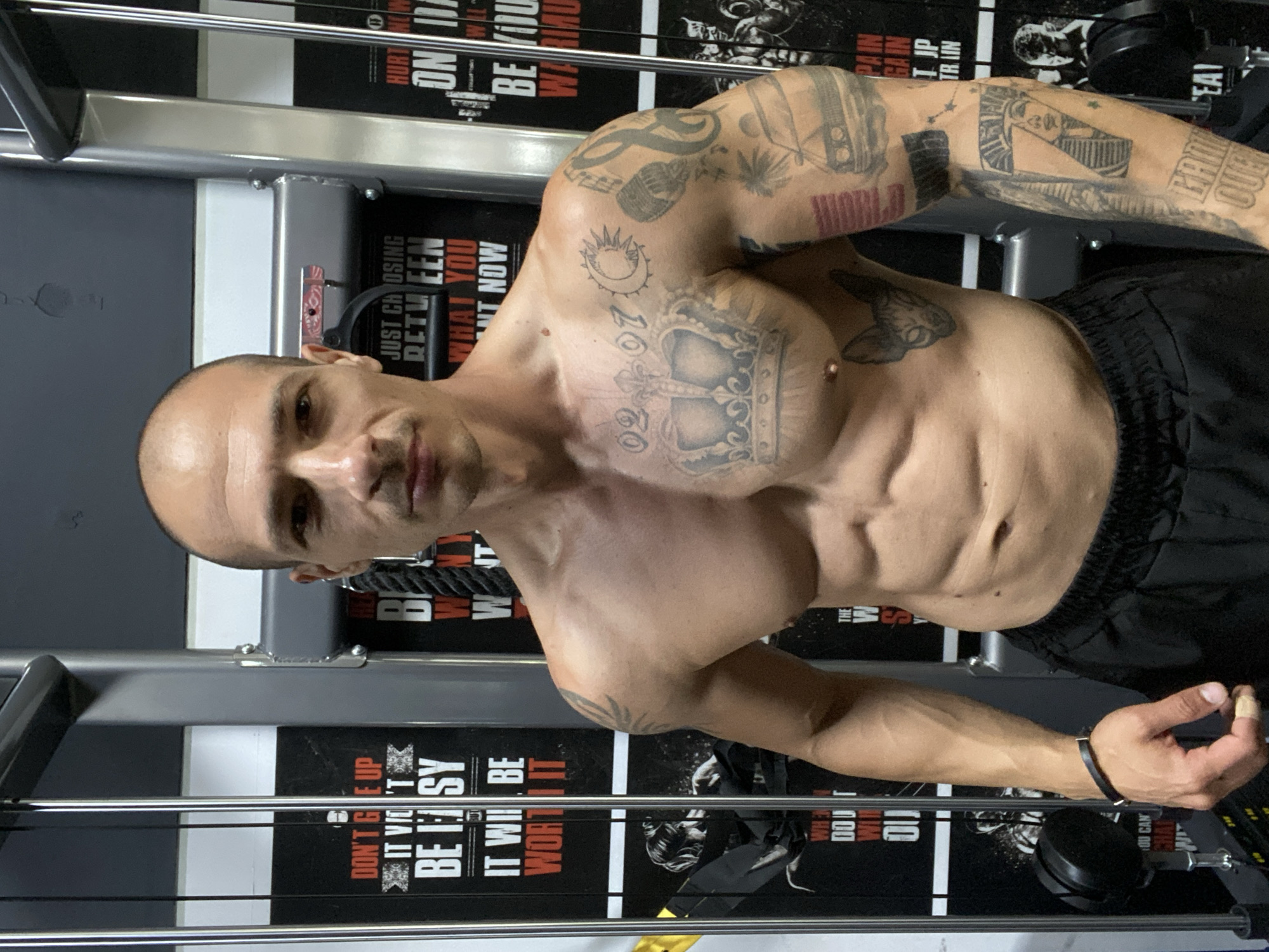 A shirtless man with a bald head and tattoos stands in a gym. He is wearing black shorts and a black bracelet on his left wrist. In the background, there are gym machines and motivational posters. The posters have text in English, such as "DON'T GIVE UP IT WON'T BE EASY IT WILL BE WORTH IT" and "JUST CHOOSING BETWEEN WHAT YOU WANT NOW". The man's body is muscular, and he has visible abs. His tattoos include a crown, a date, a sun and moon, a microphone, a treble clef, a car, and an Egyptian pharaoh. The man is looking directly at the camera with a neutral expression. The lighting suggests an indoor setting, likely a gym with artificial lights. The image appears to be a selfie taken in Pleven, Bulgaria.
