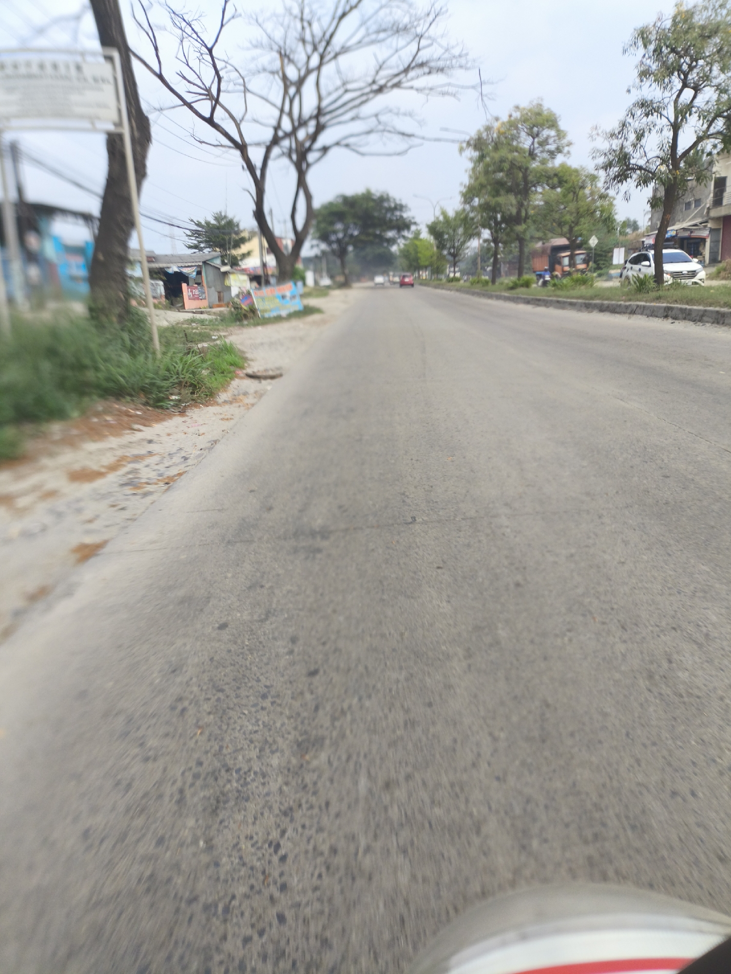 The image depicts a road scene in Cilegon, Indonesia, with an overcast sky. The perspective is low, suggesting it was taken from a vehicle. The road is paved and shows some wear and tear, with a clear lane division line faintly visible. On the left side of the road, there is a grassy verge with some scattered debris, and a signpost with writing that is mostly illegible due to blur. A few small buildings and trees line the left side of the road.
Further down the road on the left, there are more buildings, some appearing to be shops or small businesses, with tarps or awnings visible. Several vehicles, including a truck and a white car, are parked or driving on the road in the distance. The right side of the road has a raised concrete curb, and beyond that, more trees and buildings, some of which appear to be multi-story structures. The overall impression is of a semi-urban environment with moderate traffic. The motion blur in the image indicates movement, likely captured while traveling along the road.