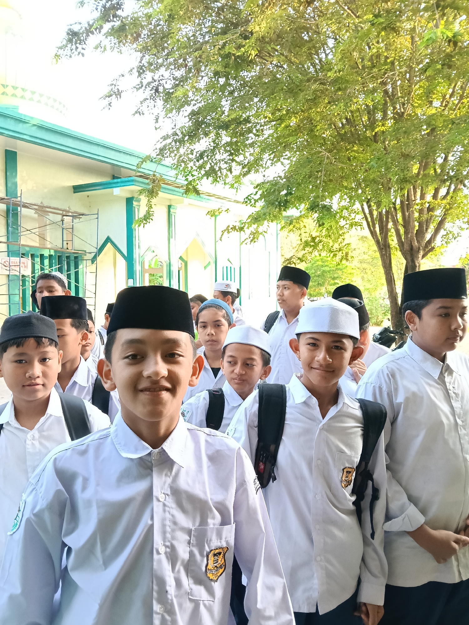 A group of male students, appearing to be of Indonesian ethnicity, are walking in a line. Most of them are wearing white collared shirts, dark backpacks, and either a black peci or a white skullcap. Some shirts have badges on the pocket. They are walking in front of a building with light-colored walls and green trim. Scaffolding is visible near the building. A large tree with green foliage is located nearby. The scene is likely in Lhokseumawe, Indonesia. The photograph is taken outdoors during daylight.