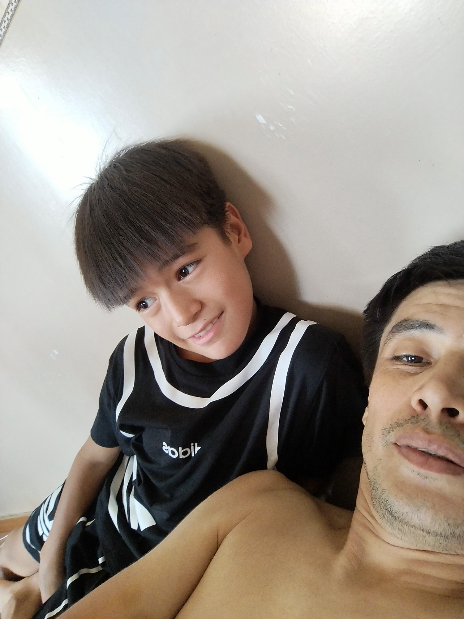 A selfie shows a young boy and an adult man indoors, likely at home, against a light-colored wall. The boy, positioned slightly to the left of the frame, has dark hair and is wearing a black t-shirt with white stripes. He is looking towards the right with a slight smile. The adult man is in the foreground on the right, with dark hair and stubble. He is also looking towards the camera and appears to be smiling faintly. The scene is casually posed, suggesting a relaxed moment between the two individuals. The lighting indicates it is daytime. The text "apbil" is visible on the boy's t-shirt.