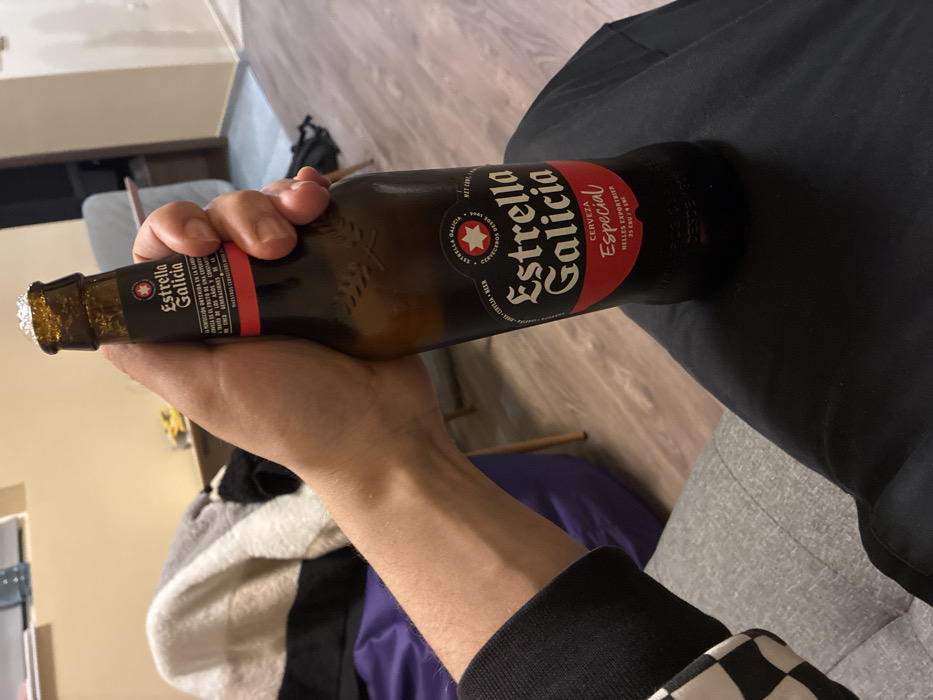 This image captures a close-up, first-person perspective from an indoor residential setting located in Sofia, Bulgaria. A right forearm and hand are prominently featured, gripping a 330ml brown glass bottle of "Estrella Galicia Especial Cerveza." The bottle exhibits condensation on its surface, indicating it is cold, and its cap is removed, with a small amount of foam visible at the opening. The person's arm is partially covered by a dark sleeve with a distinct white and black checkered pattern visible at the cuff. The individual appears to be seated on a grey upholstered sofa, a portion of which is visible in the lower left. The background reveals light brown wooden laminate flooring, a purple beanbag chair positioned to the left, and a light-colored blanket with white, grey, and black stripes draped over an object further back. A plain, light-colored wall and a partial view of a light-toned table are also discernible in the background. No other individuals are present or interacting within the visible frame.