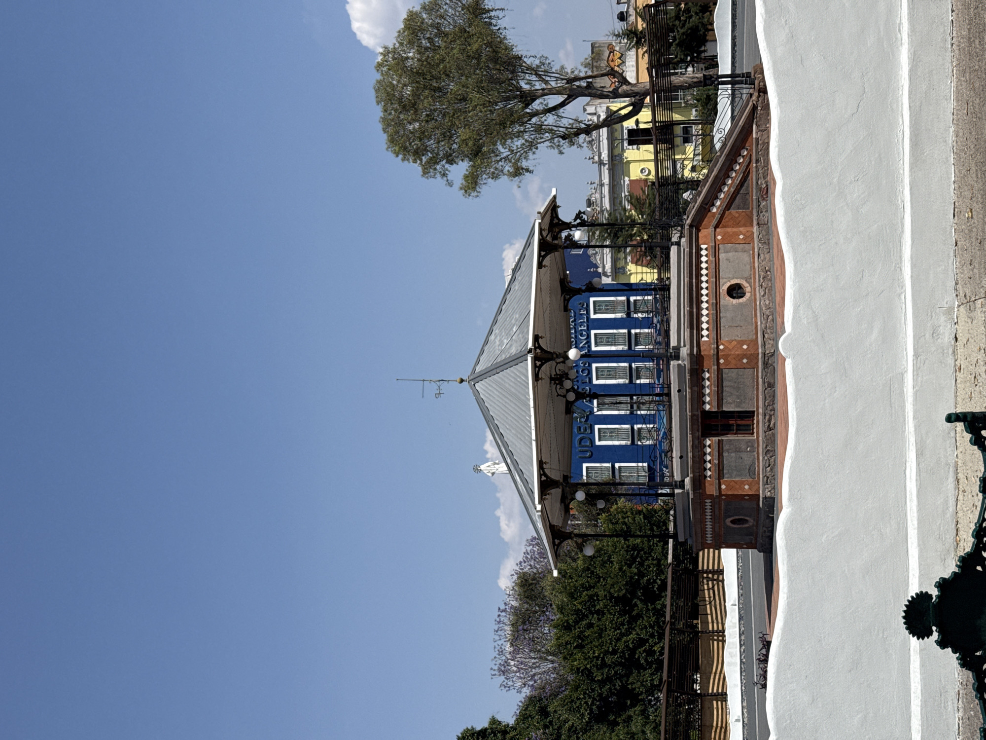 The image shows the kiosk (El Kiosco) in the Zócalo (main plaza) of Puebla, Mexico, set against a clear blue sky. The kiosk features a blue façade with white trim, windows, and lettering, and a silver-colored, multi-tiered roof. Trees with green and lavender foliage surround the structure. In the background, there are yellow and tan buildings. A portion of a white wall with an ornate green metal fixture is visible on the right.