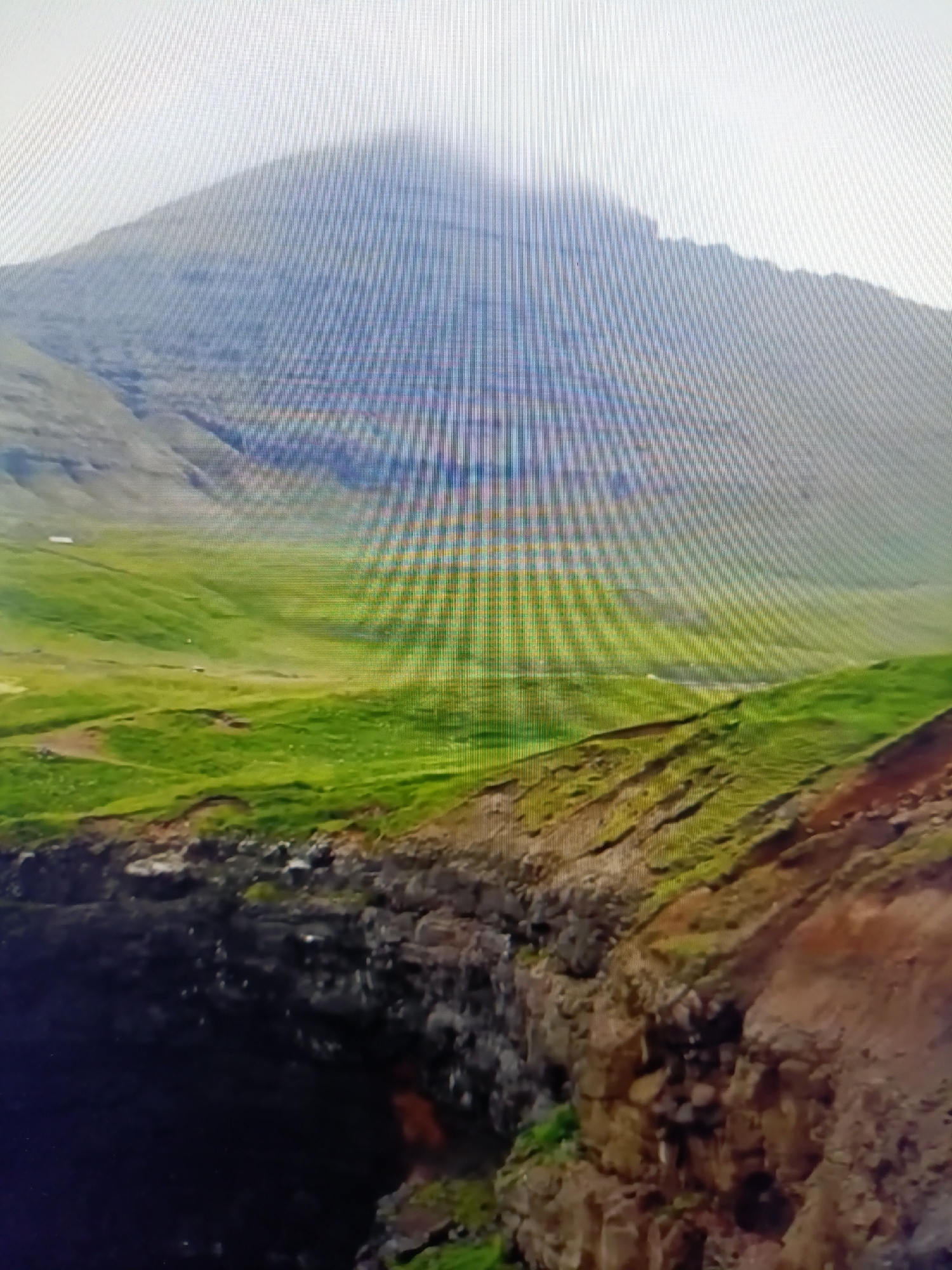 This is a photograph of a dramatic coastal cliff scene. In the foreground, rugged, dark rocks form the edge of a steep drop-off into a dark abyss. Above the cliff, rolling green hills stretch into the background, leading to a large, mist-shrouded mountain. The sky is overcast and bright. There are no people or identifiable objects in the image to suggest a specific location within Netanya, Israel. The image appears to be a landscape photograph of a natural environment.