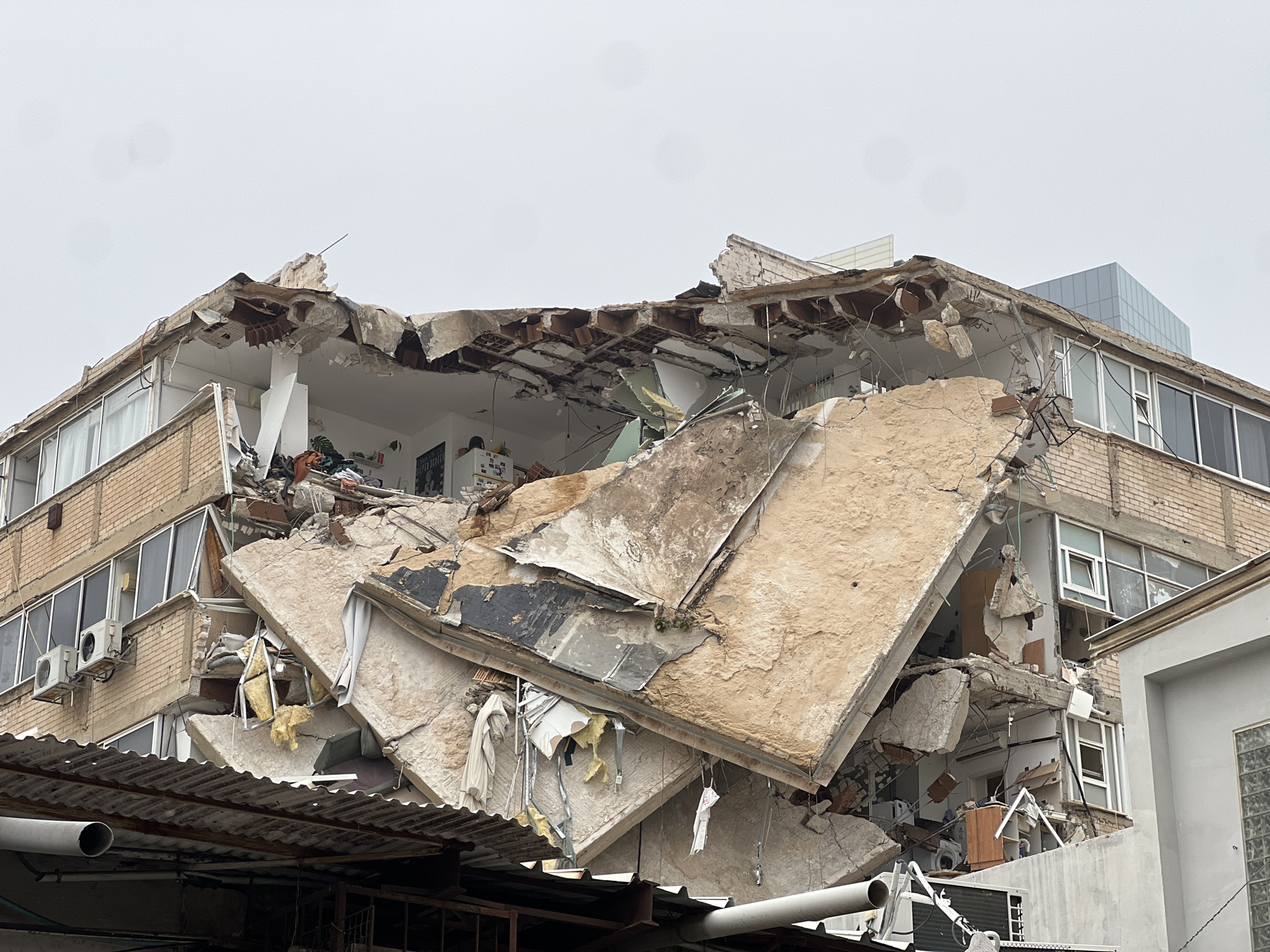 The image shows a partially collapsed building in what appears to be an urban area. The facade is damaged, revealing the interior of the structure. Concrete debris and exposed building materials are visible. The building has a brick exterior on the intact sections. There are signs of interior furnishings and personal belongings amidst the wreckage. Outside the building debris is on top of other structures like an awning or corrugated metal structure. The sky is overcast. The city and country are unknown, but the prompt suggests Tel-Aviv, Israel. There is no sign of people in the scene.