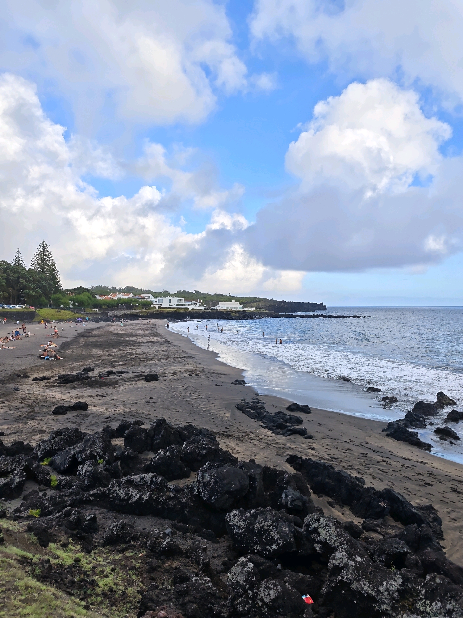 This is a photograph of a black sand beach in Ponta Delgada, Portugal. The beach is populated by numerous people sunbathing, walking, and swimming in the ocean. The water is blue and calm, with small waves lapping at the shore. In the background, there are several white buildings and lush green trees. The sky is partly cloudy with patches of blue visible. The foreground of the image features dark, volcanic rocks scattered across the sand.
