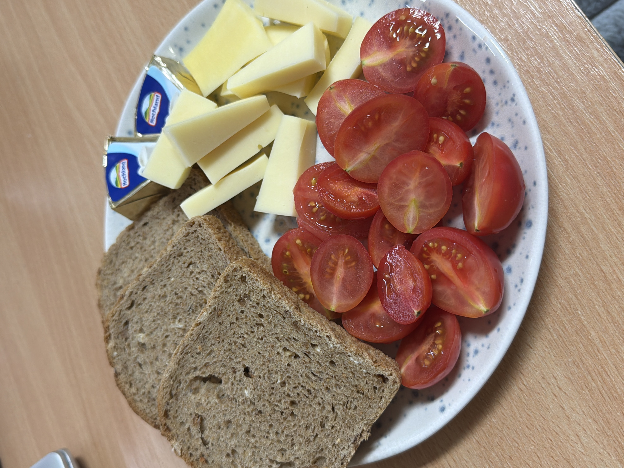 This media file captures a close-up overhead view of a prepared meal or snack served on a white plate with blue speckles. The plate is positioned on a light-colored wooden table, suggesting an indoor setting in Sofia, Bulgaria.
The meal consists of several food items. On the left side of the plate, there are numerous pieces of light yellow cheese, cut into rectangular or wedge shapes. Adjacent to these, two small, triangular portions of foil-wrapped cheese are visible, clearly displaying the brand name "Hochland" on their blue and gold packaging. The right side of the plate is filled with a generous serving of bright red cherry tomatoes, all cut in half, revealing their juicy interiors and seeds. Below the plate, extending from the bottom left, are three slices of dark brown, rustic-looking bread, likely rye or whole grain.
The overall presentation is simple and fresh. No people are visible, and no specific activity is taking place other than the arrangement of the food. The lighting appears natural, suggesting daytime.