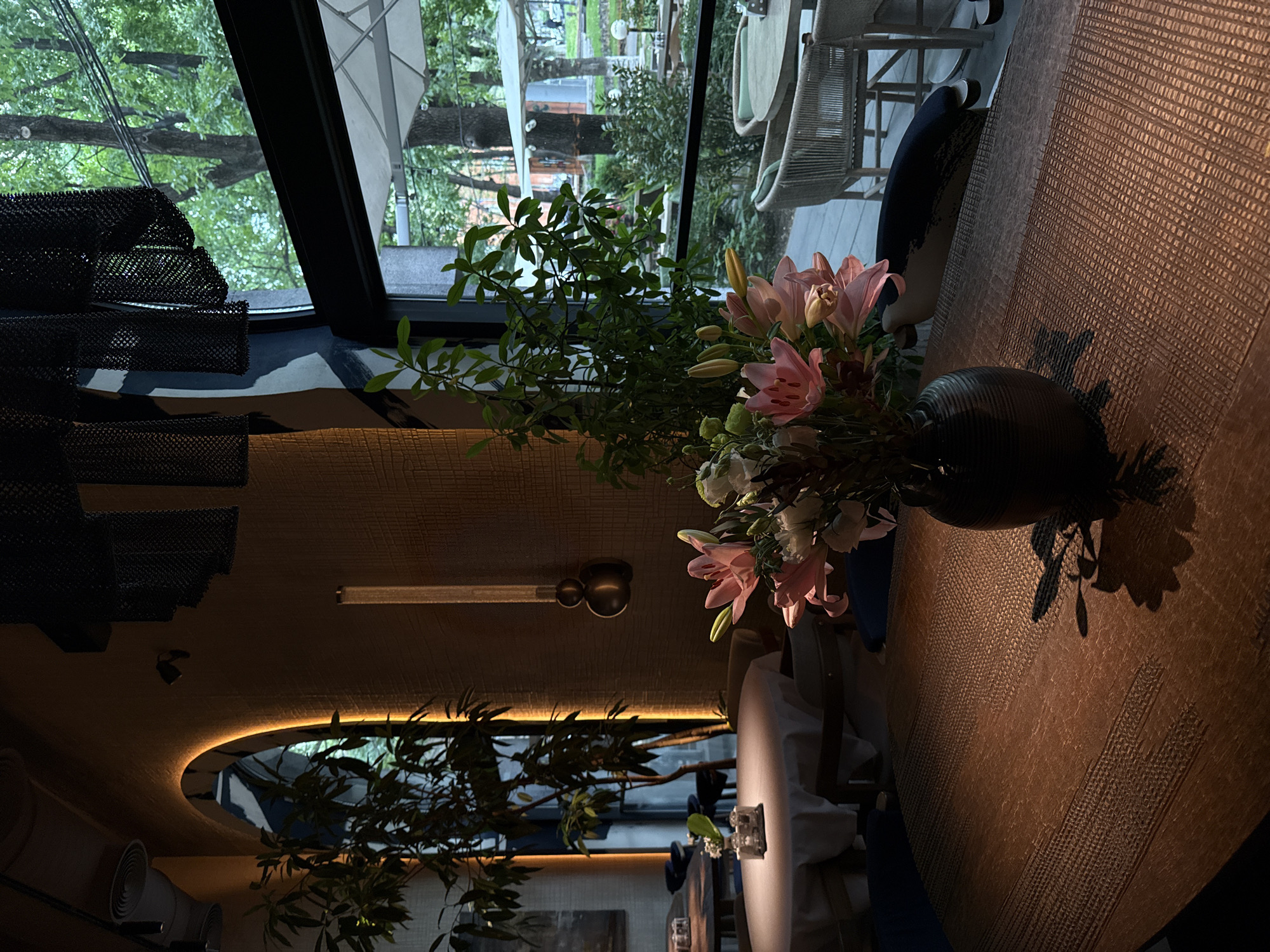 The image depicts the interior of a sophisticated restaurant or cafe in Yerevan, Armenia, featuring a large window that offers a view of an outdoor garden or patio. In the immediate foreground, a striking bouquet of fresh flowers, predominantly composed of pink lilies, green foliage, and some white blooms, is arranged in a dark, textured vase positioned on a table. The table itself has a distinct woven or textured surface, casting subtle shadows from the flowers.
The interior space is characterized by dim, ambient lighting. Warm, indirect illumination emanates from an arched ceiling feature, highlighting textured wall or ceiling panels. On the left side of the frame, black mesh or woven decorative elements add to the contemporary aesthetic. Through the expansive window, lush green trees and other foliage are visible, suggesting a vibrant outdoor environment. Hints of outdoor furniture, including a white umbrella and light-colored wicker chairs, can be seen beyond the window.
The natural light filtering in from the outside contrasts with the subdued lighting indoors, creating a serene and intimate atmosphere. No people are present in the scene, and no specific activity is taking place, but the setting is clearly prepared for dining or relaxation. No text is visible within the image.