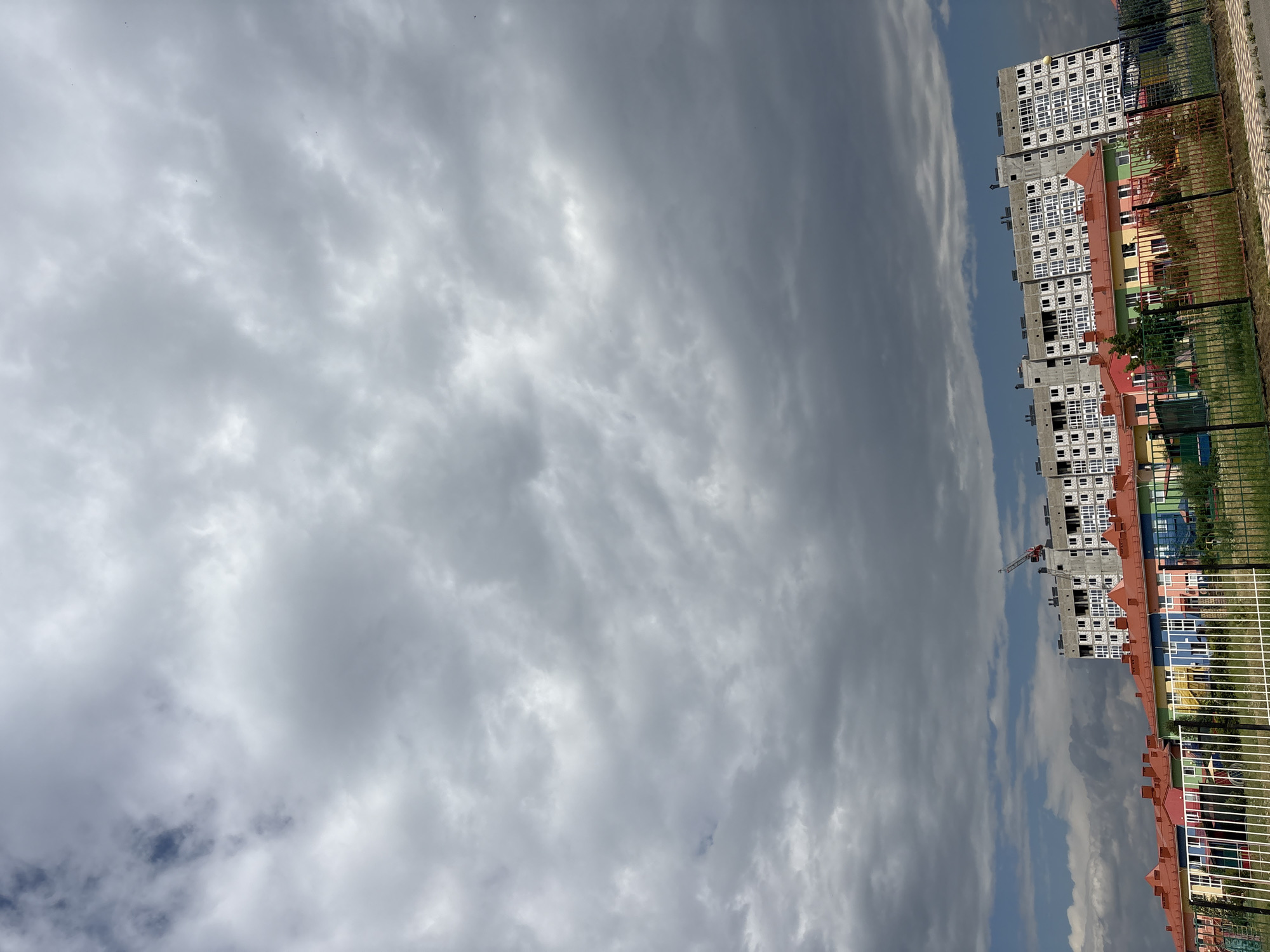 The image shows a construction site with a building under construction in Mikhaylovsk, Russia. In the foreground, there are several brightly colored, single-story buildings with red roofs, resembling a kindergarten or daycare facility. Behind these, a large, multi-story building is under construction, appearing to be made of concrete with many windows. A construction crane is visible above this building. The sky is filled with dramatic, dark gray clouds, suggesting overcast or impending rain. A black metal fence runs along the bottom of the image, separating the foreground from the buildings. The scene appears to be an outdoor environment on a cloudy day.