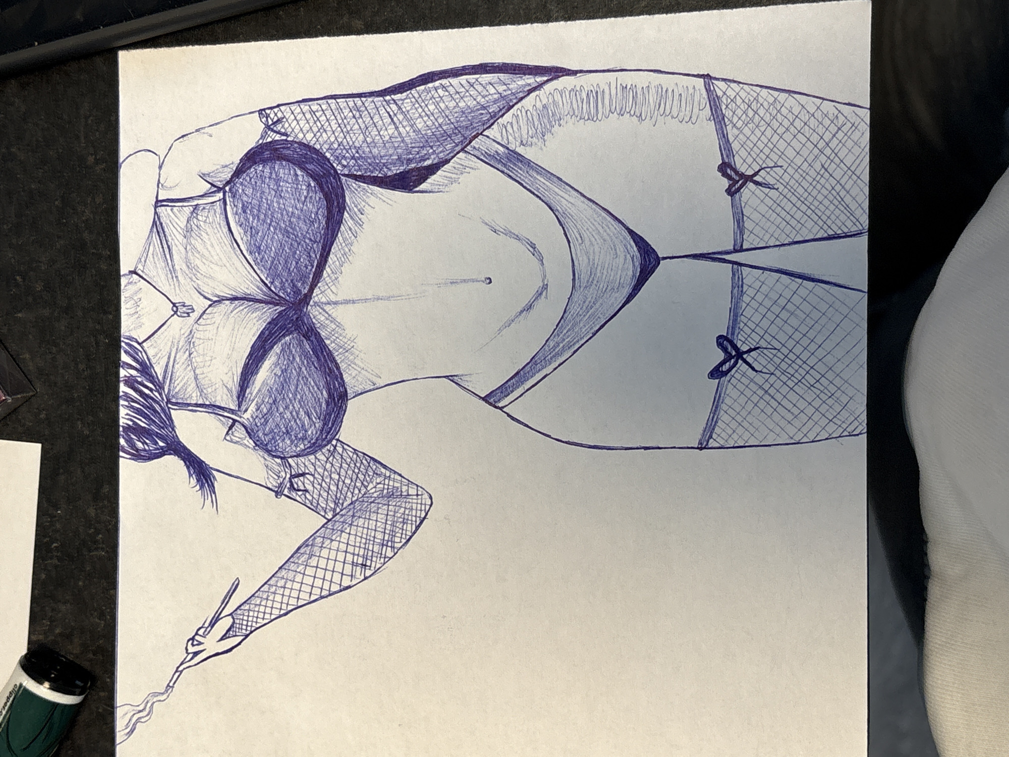 The image is a pen drawing of a woman in lingerie, including a bra, panties, and fishnet stockings with decorative bows. She is holding a long, thin object, possibly a cigarette or a paintbrush, with wisps of smoke or paint rising from it. The woman is depicted in a reclining position. The drawing is on a piece of paper, which is placed on a dark-colored surface. A green and white marker or pen is visible in the lower left corner of the image. A white object is visible on the right side.