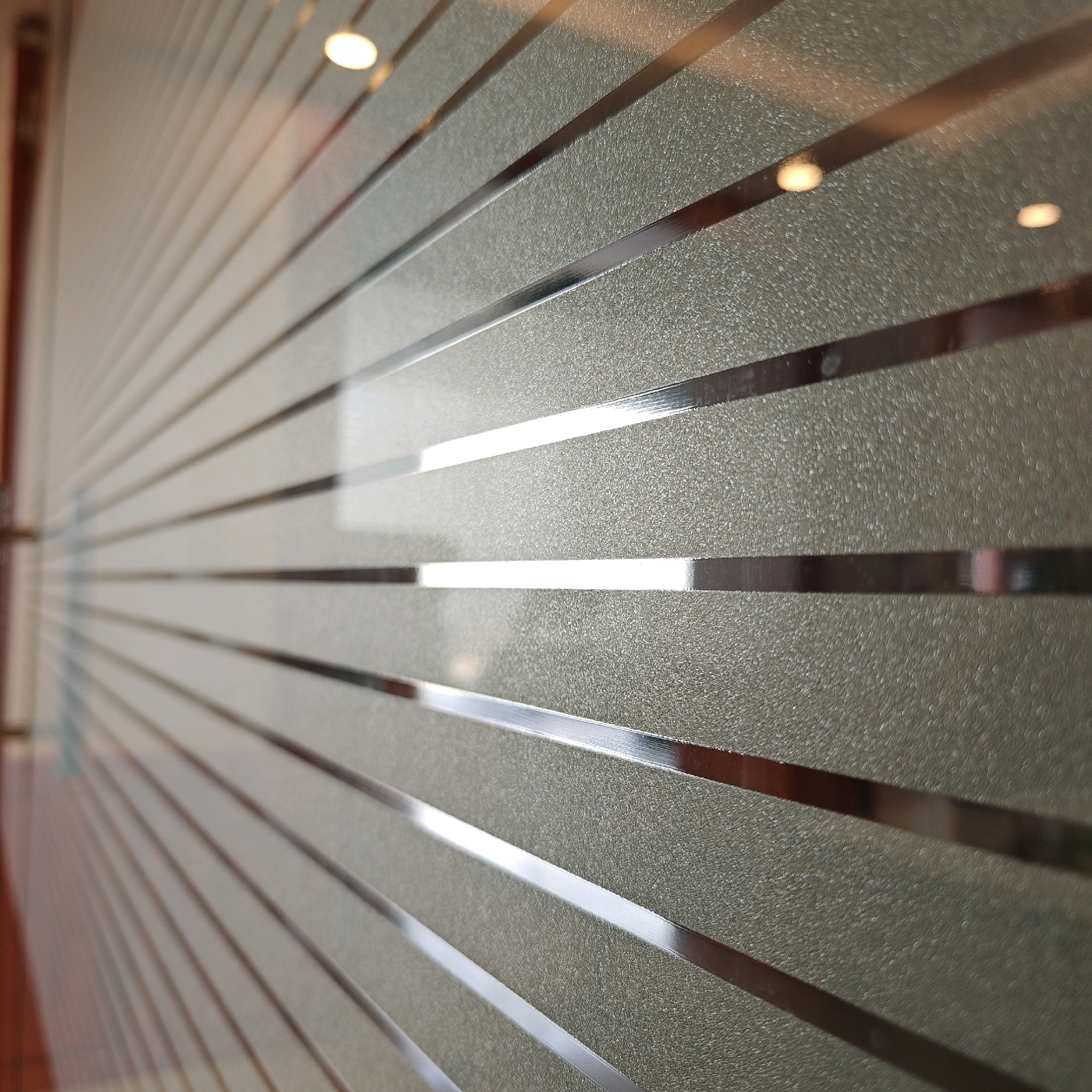 The image shows a close-up of a frosted glass partition with horizontal metallic stripes. The partition is angled, with the foreground in sharp focus and the background slightly blurred, suggesting a shallow depth of field. Light reflects off the metallic stripes and also shines through the frosted glass, creating bright spots and a speckled texture. On the far left, a hint of a wooden frame and a door handle can be seen, suggesting this is an interior partition, possibly in a building or office. The overall impression is one of modern interior design. The location in Gulab Bagh, India, is not visually discernible from the image itself.