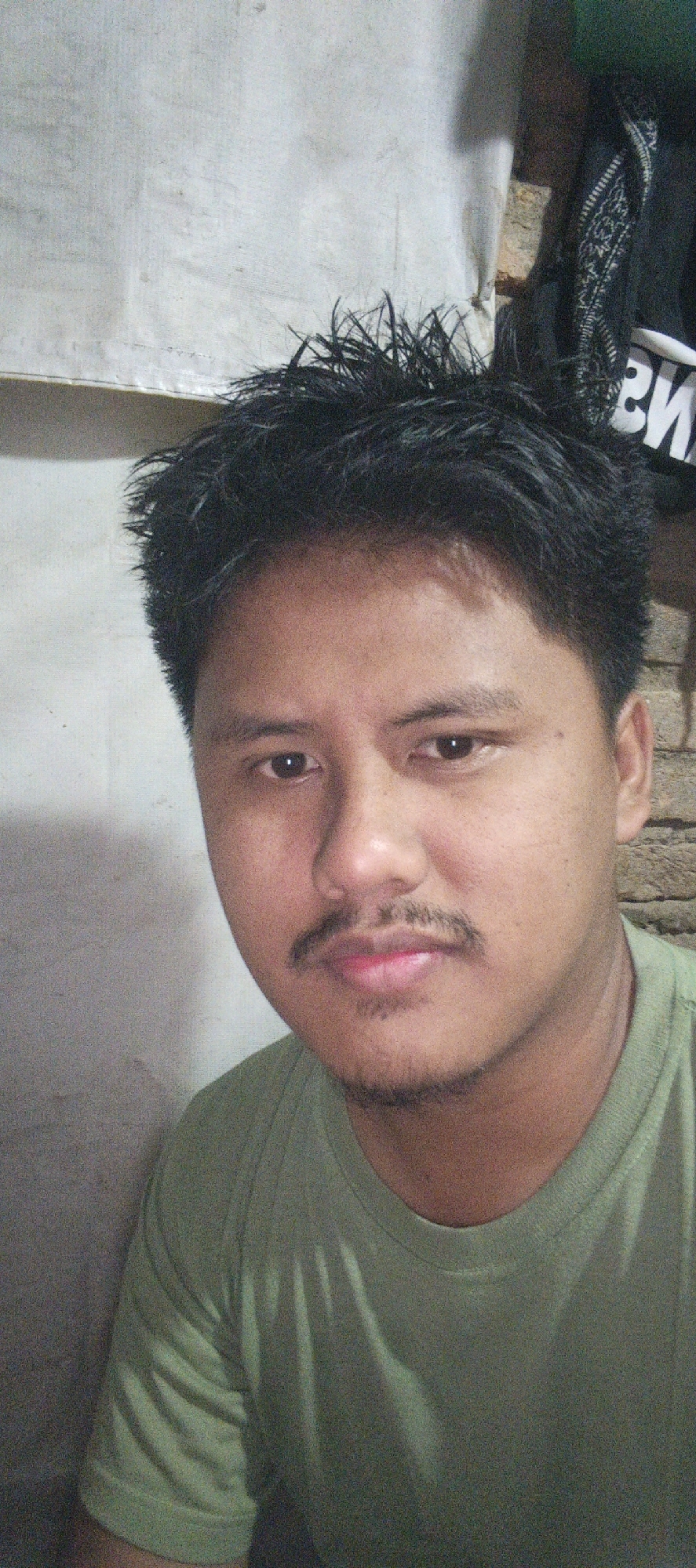 This is a portrait of a young man indoors, possibly in Kemuning, Indonesia. He is looking directly at the camera with a neutral expression. He has dark, spiky hair and a mustache and a small patch of stubble on his chin. He is wearing a green t-shirt. The background is a textured, light-colored wall with some discoloration and a dark object with a patterned fabric in the upper right corner. The lighting is somewhat dim, casting shadows on the man's face and the wall.