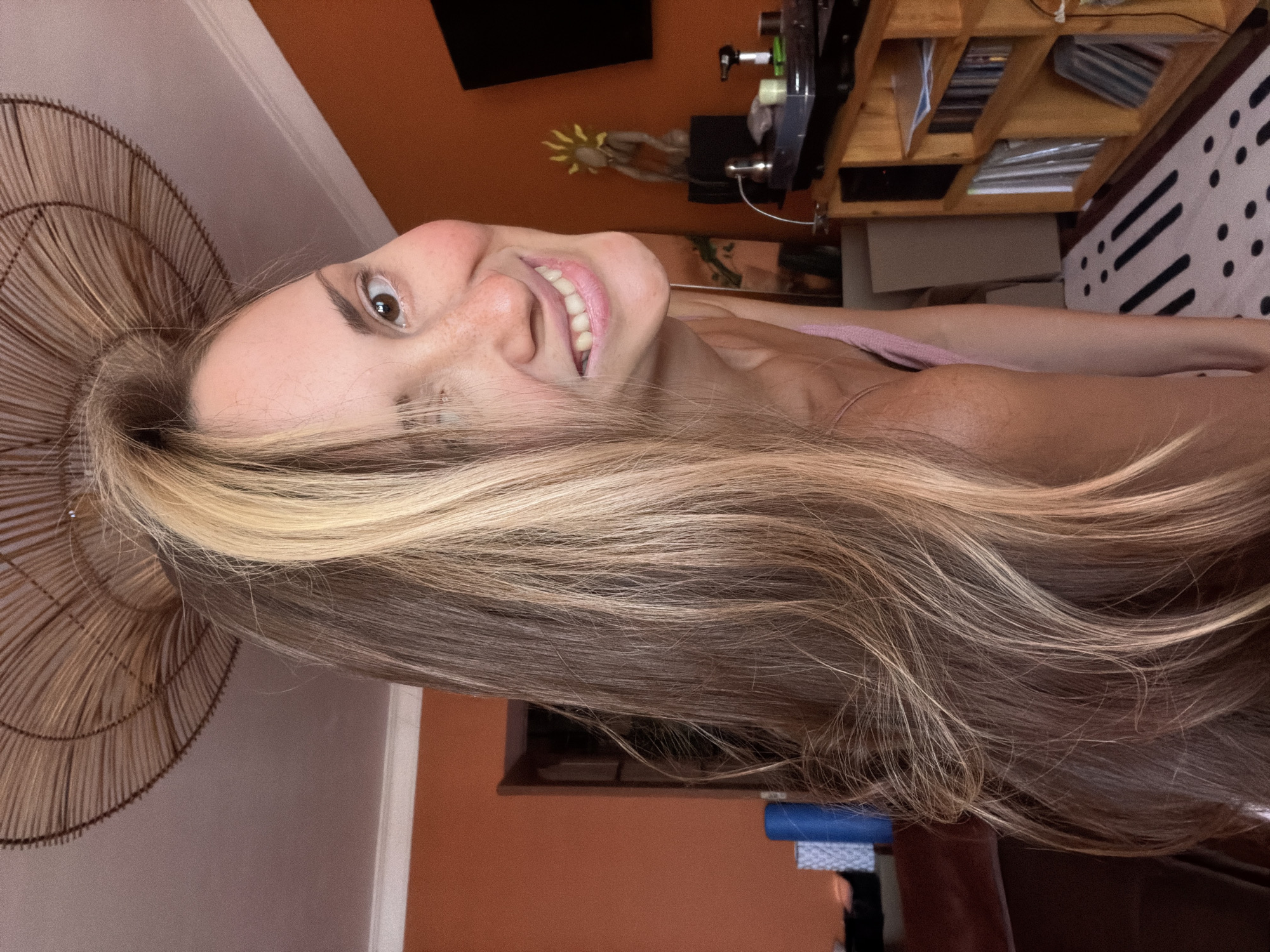 The media file is a close-up, rotated selfie of a woman with long, light brown and blonde hair. She is looking up and smiling with her mouth slightly open, showing her teeth. Freckles are visible on her nose and cheeks. Her left eye is looking directly at the camera, while her right eye is partially obscured by her hair. She is wearing a light pink top.
The background is a room with an orange wall. To the left, there is a woven, circular object. Behind her head and to the right, there is a wooden shelf unit filled with various items, including books or binders. A black rectangle is visible on the wall above the shelf. A decorative sunflower-like object is also visible on the orange wall. A black and white patterned textile is draped over a surface to the far right.
The lighting suggests it is daytime, possibly indoors with artificial light. The overall mood appears happy and relaxed. There is no visible text in the image. The location context is Khimki, Russia.