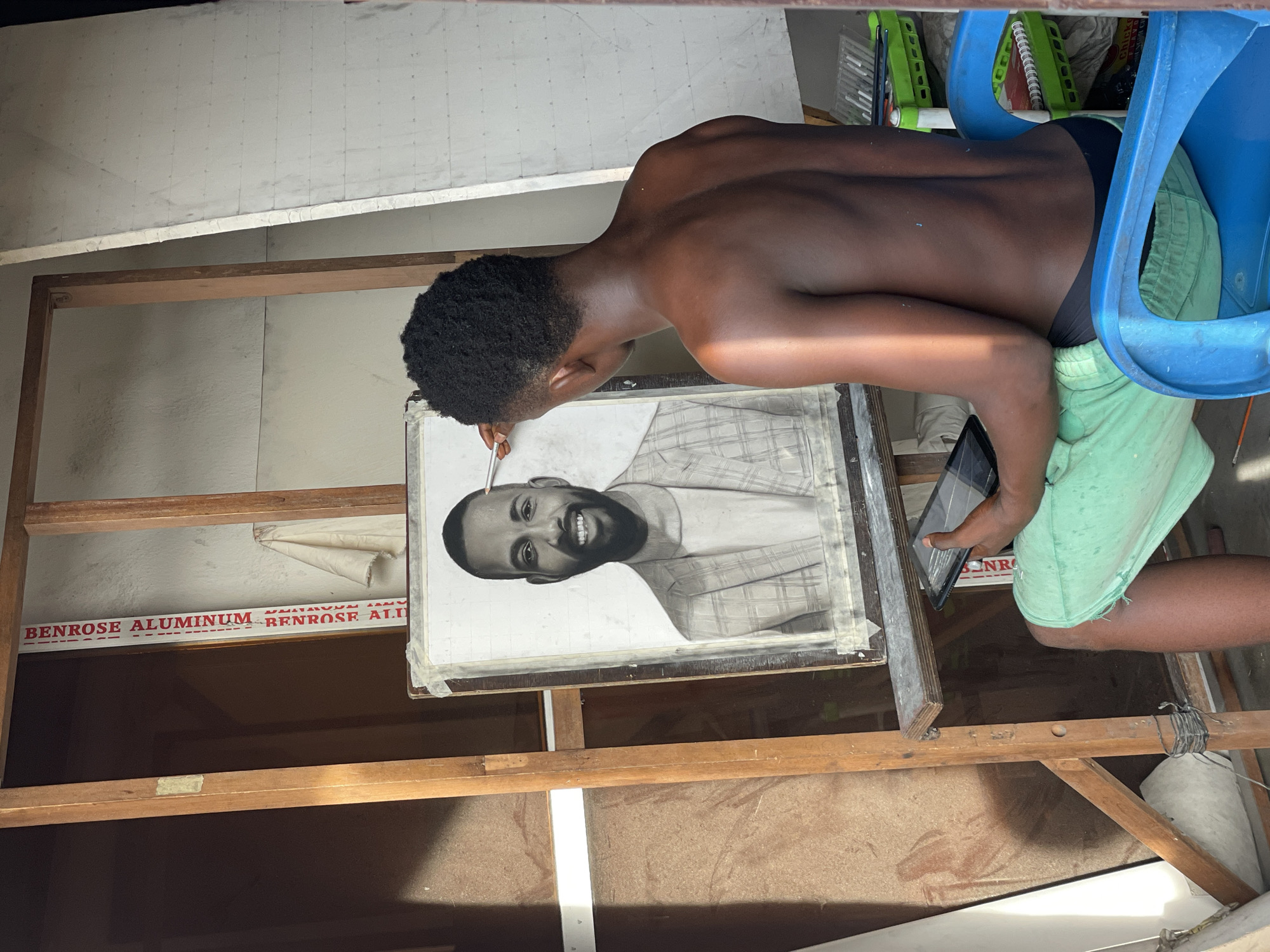A young, dark-skinned man with short, dark hair sits on a blue plastic chair, holding a tablet in his left hand and a paintbrush in his right, as he works on a portrait in a frame. The portrait appears to be a black and white rendering of a man with a beard and a plaid shirt. The artwork is on an easel made of wooden beams. The backdrop includes a wall that is partially covered with what seems to be an aluminum sign marked "BENROSE ALUMINUM." The man is shirtless and wearing light green shorts. The setting appears to be a simple, outdoor workshop environment, possibly in Onitsha, Nigeria.
