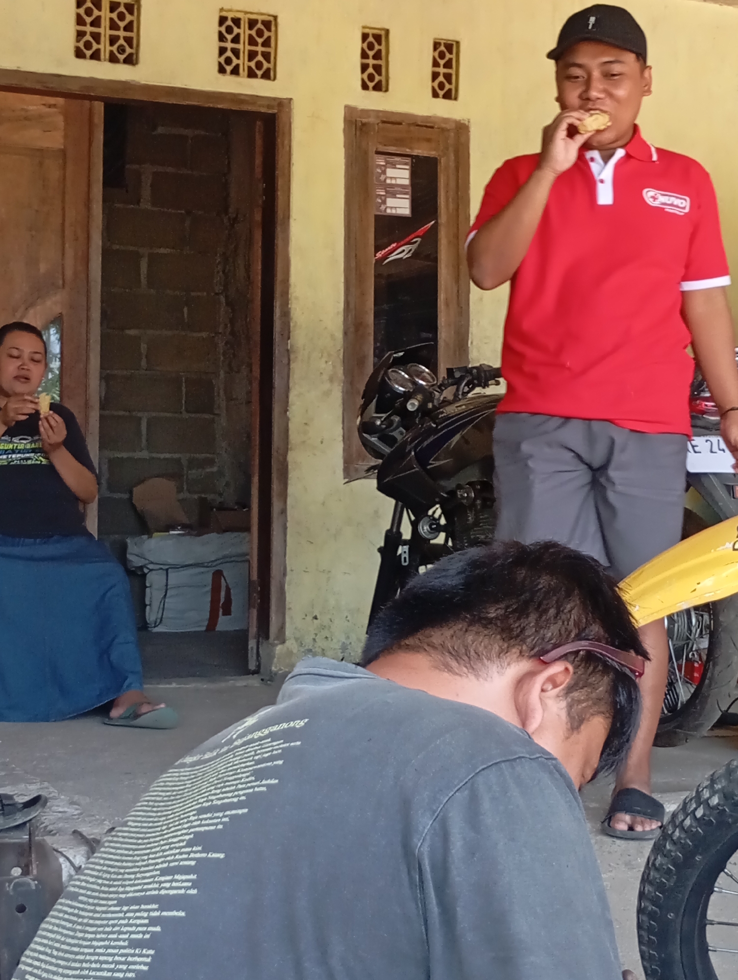 In Ketepung, Indonesia, a man wearing a red polo shirt and a black cap is standing, holding a piece of food to his mouth. To his left, a woman in a dark t-shirt and a blue skirt sits. Behind them, a motorcycle is parked. In the foreground, a person wearing a dark grey t-shirt is seated, their back to the camera. The scene appears to be outdoors, possibly in front of a building with yellow walls and decorative ventilation bricks. The overall environment suggests a casual, possibly domestic setting.