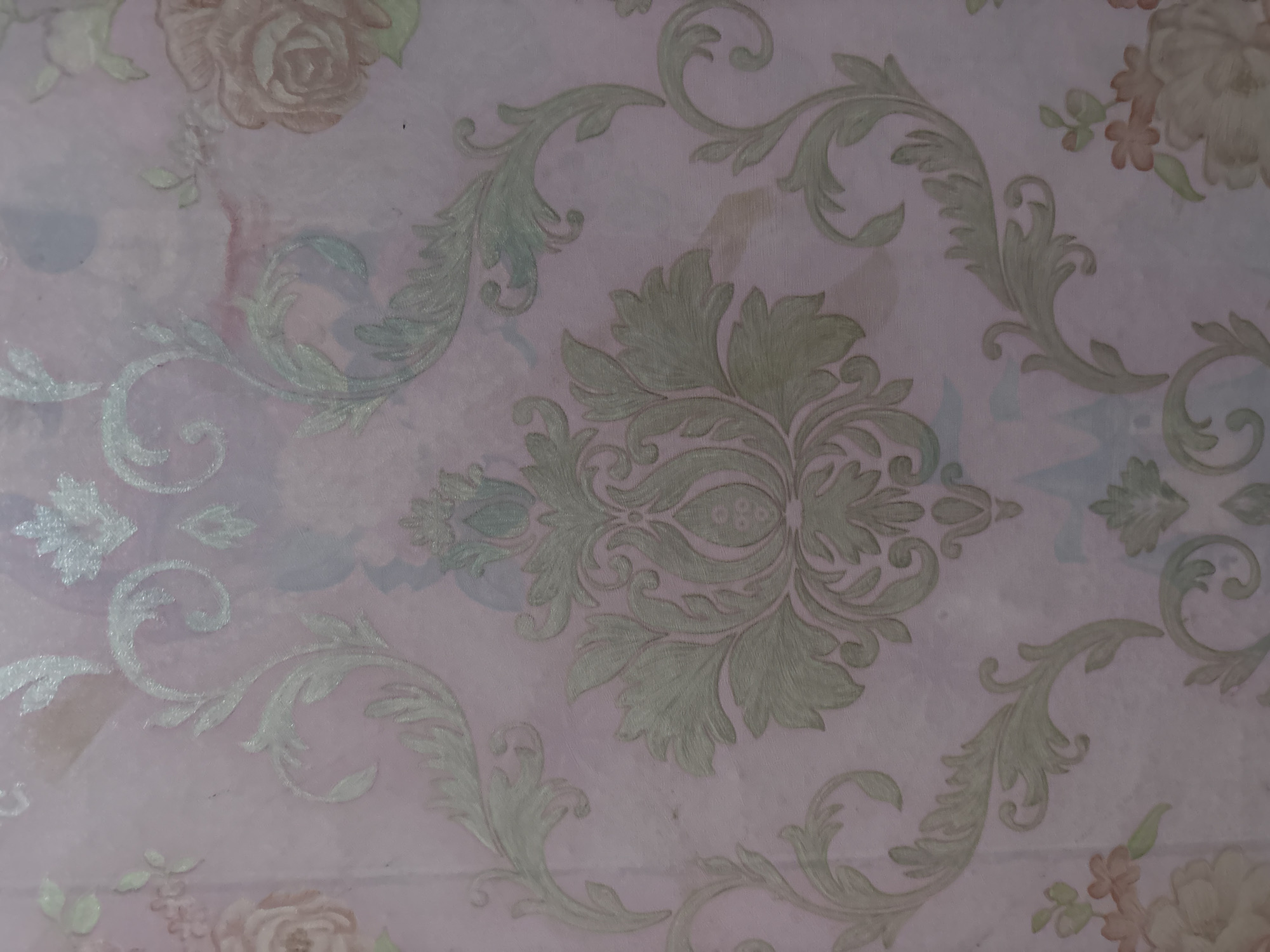 The image shows a close-up view of floral damask wallpaper with a pale pink background. The pattern features an ornate damask design in a muted green and light silver hue, interspersed with stylized floral motifs and swirling vines. The lighting is soft and diffused, creating a slightly textured appearance. There are no people or discernible objects from the city of Danawinangun, Indonesia, present in the image.