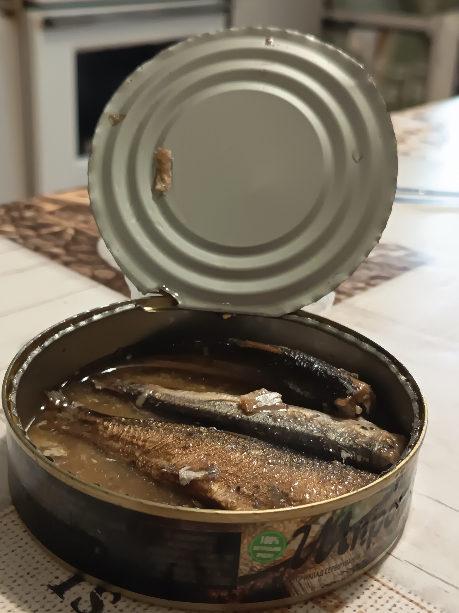 A high-angle, close-up shot shows an open tin of sardines. The sardines are packed tightly in oil and are layered on top of each other. The lid of the tin is partially open and rests at an angle, revealing the contents. The tin has a black label with some text and graphics. The scene appears to be indoors, with a light-colored wooden surface visible beneath the tin and a patterned placemat. The background is blurred but suggests a kitchen environment. There are no people or animals in the image. The location in the city of Izmail, Ukraine is not specified or evident in the image.