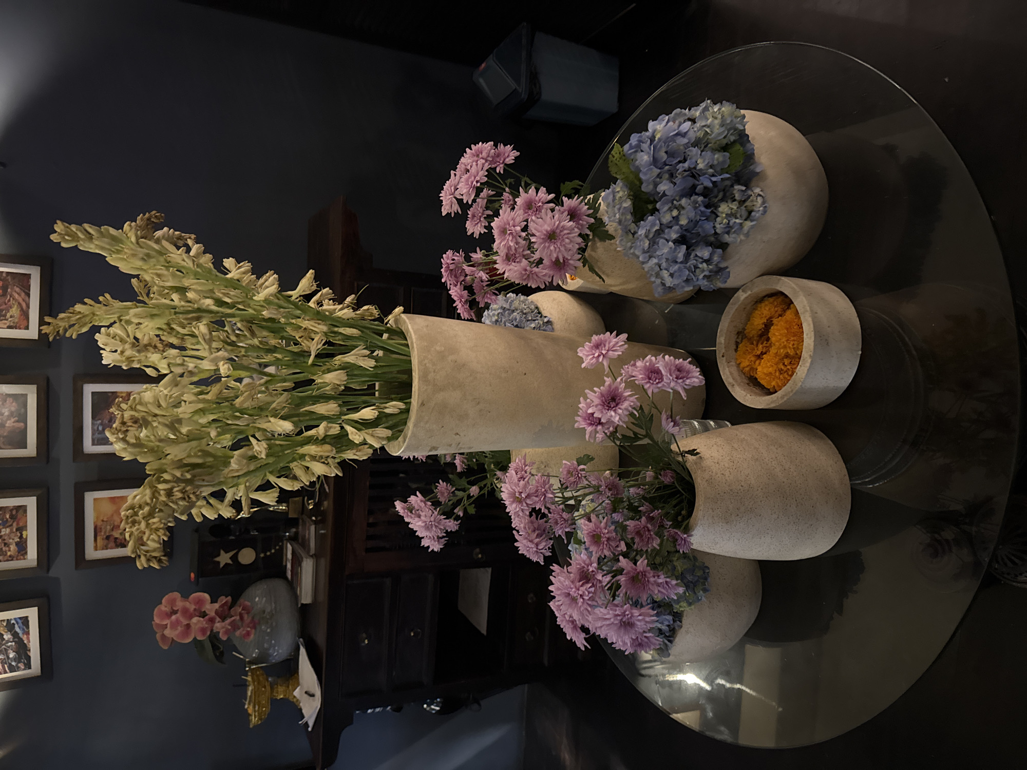This image depicts an indoor scene, likely a reception area or living room, with a table adorned with floral arrangements. The table is round and appears to be made of dark glass.
Dominating the center of the table is a tall, pale beige vase filled with dried, pale yellow tubular flowers. To its left and slightly in front, a smaller, similarly colored vase holds a bouquet of vibrant pink chrysanthemums. Behind the pink flowers, a pale blue hydrangea arrangement is partially visible in another beige vase. To the right of the central vase, a small, round, concrete-like container holds bright orange marigold petals.
In the background, a dark wooden credenza with drawers and slatted doors is visible. Above the credenza, several framed pictures hang on a dark blue wall. To the left of the credenza, a pink orchid in a light blue, round vase sits on the surface. A golden decorative object rests on a white napkin to the far left. A dark blue trash can is visible to the far right in the background.
The lighting is dim, creating a moody atmosphere with soft highlights and shadows. The image is composed from a medium-low angle, looking slightly up towards the floral arrangements. There are no people visible in the image. The overall aesthetic suggests a sophisticated and tranquil environment.
This scene is consistent with decorative elements found in various establishments and residences in the city of North Kuta, Indonesia, known for its hospitality sector and cultural influences.