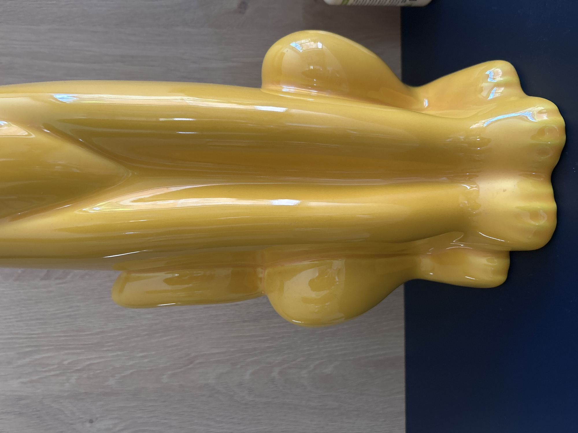 The image shows a yellow, glossy, ceramic piece on a surface. Part of the object sits on what appears to be a light-colored wooden floor, while the other part rests on a dark blue surface. The shapes of the ceramic piece are rounded. Part of a white and green object is visible in the upper-center of the image.