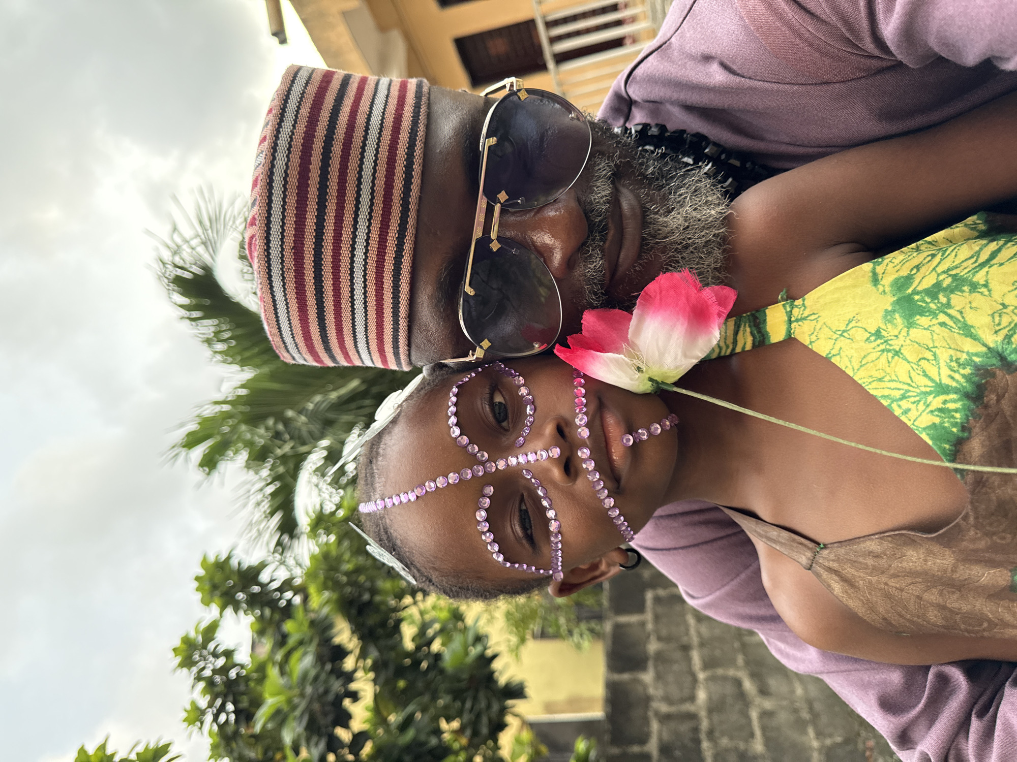 An outdoor portrait captured in Ado, Nigeria, features an adult male embracing a child. The male subject, positioned on the right, possesses dark skin, a prominent grey beard, and wears a horizontally striped cap composed of red, black, white, and pinkish-brown bands. He also wears dark aviator-style sunglasses with gold accents, a long-sleeved purple garment, and a black beaded necklace. The child, situated in front and slightly to the left, has dark skin and directly faces the viewer. Their face is decorated with numerous small, circular, light purple rhinestones arranged in a symmetrical pattern, and silver butterfly-shaped embellishments are present in their braided hair near the temples. A dark hoop earring is visible on the child's left ear. The child wears a halter-style top, brown with a leaf-like pattern on the left, and yellow-green with a tie-dye or patterned design on the right. A single artificial flower with a pink and white blossom and a green stem is held by the child, positioned between the two individuals. The background consists of dense green foliage, including broad-leafed trees and palm fronds, against an overcast sky. Portions of a light yellow building with dark windows are visible in the upper right background, and a grey cinder block wall is present in the lower left.