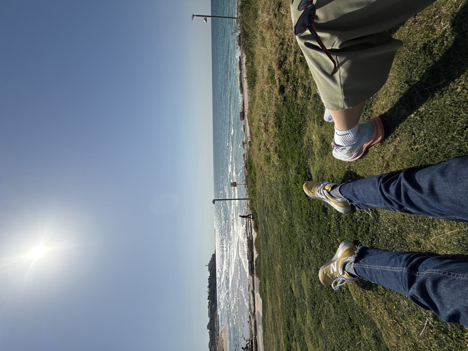 The image presents a bright, sunlit view from a low angle, depicting the extended legs of two individuals reclining on green grass. The person on the left wears blue denim jeans and yellow-and-grey athletic shoes, while the person on the right wears olive green wide-leg pants and multi-colored (light blue, pink, white) athletic shoes. Dark-framed sunglasses rest on the right individual's pants.
The scene overlooks the Mediterranean Sea, specifically along the coastline of Tel Aviv, Israel, looking south towards Old Jaffa. In the midground, a paved pathway separates the grassy area from a sandy beach and the sea, which shows moderate waves and significant sun glare. The historic city of Old Jaffa (Yafo) is clearly visible on a headland in the background on the far left, characterized by its distinctive buildings and a lighthouse structure.
A lone individual walks along the paved path in the midground. Two modern, dark-colored streetlights are positioned near the path, one on the left and one on the right. A red-and-white warning sign on a pole is also visible near the path on the left. The sky is clear and bright blue, with the sun prominently situated in the upper-left quadrant, casting a visible lens flare.