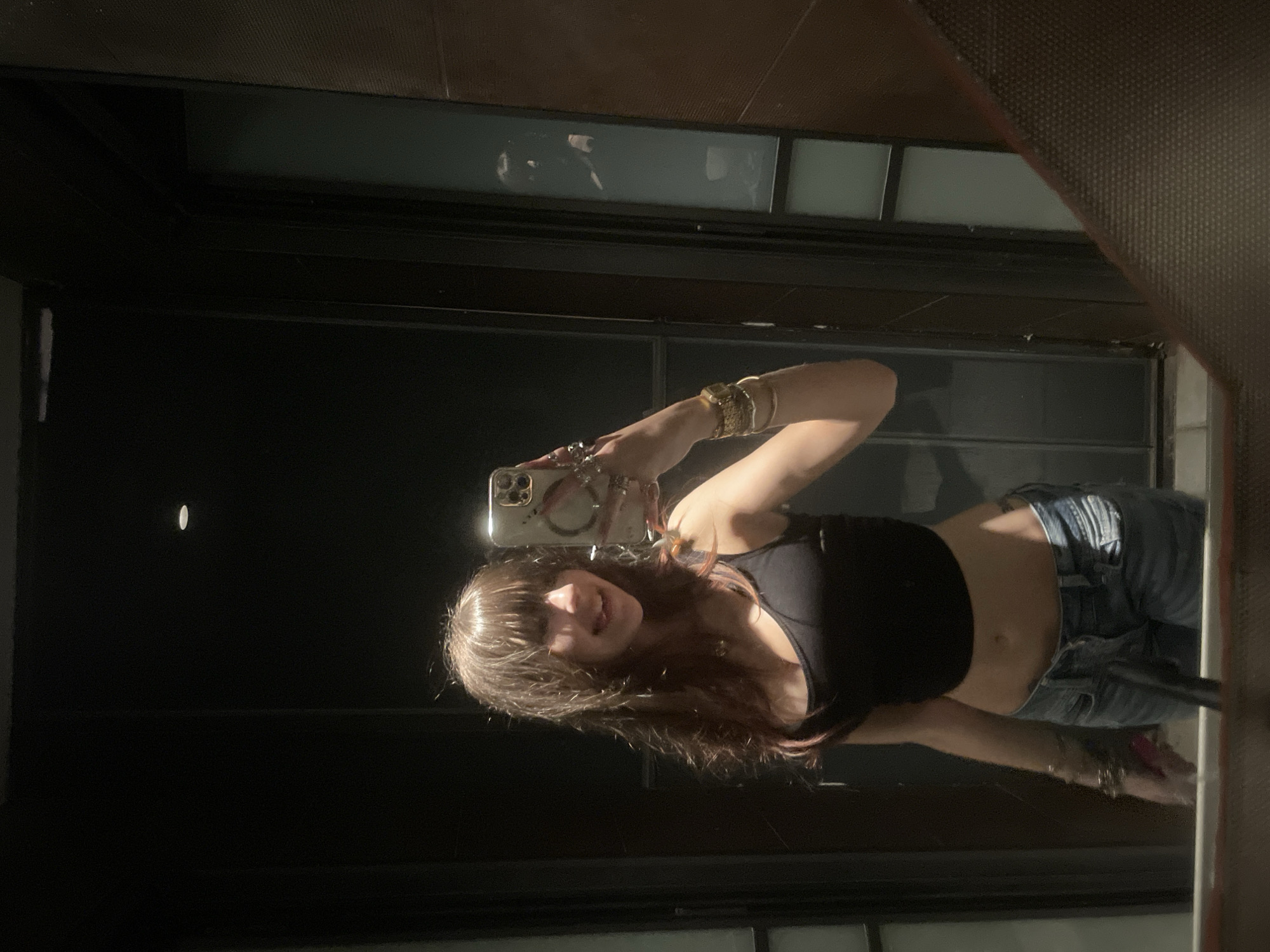 A young woman is taking a selfie in a mirror. She is wearing a black crop top and blue jeans. The mirror appears to be located in a building with a dark background with a window pane. There is no explicit information available to confirm the location is in Tel-Aviv, Israel.