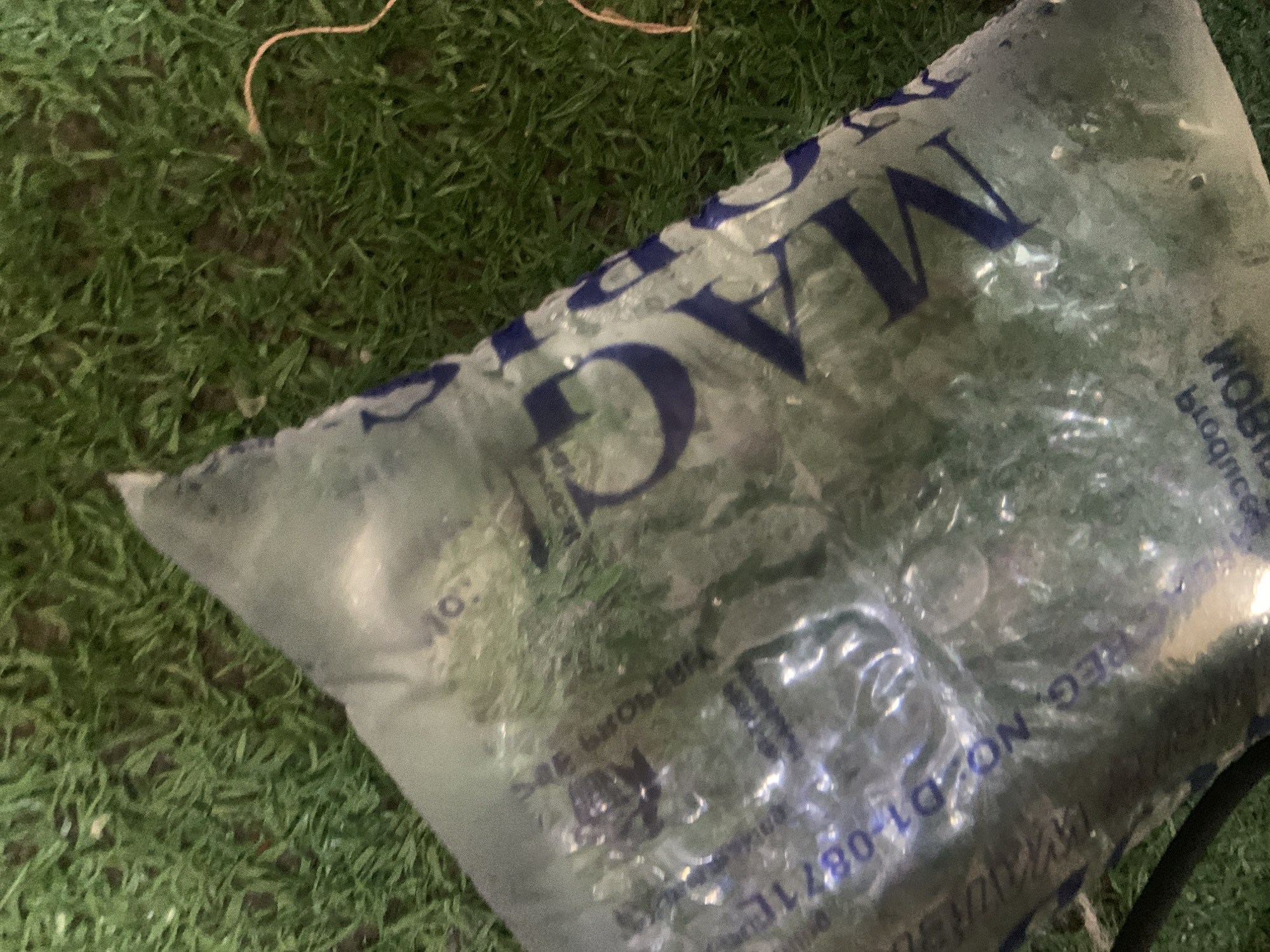 In the city of Jauro Yinu, Nigeria, a clear plastic bag filled with green matter, possibly vegetation or ice, lies on a surface resembling artificial grass. The bag has blue lettering on it, some of which is legible, including "REG. NO. D1-08711" and partial words like "VATIC" and "NOBI." The lighting in the image suggests it was taken in low-light conditions.