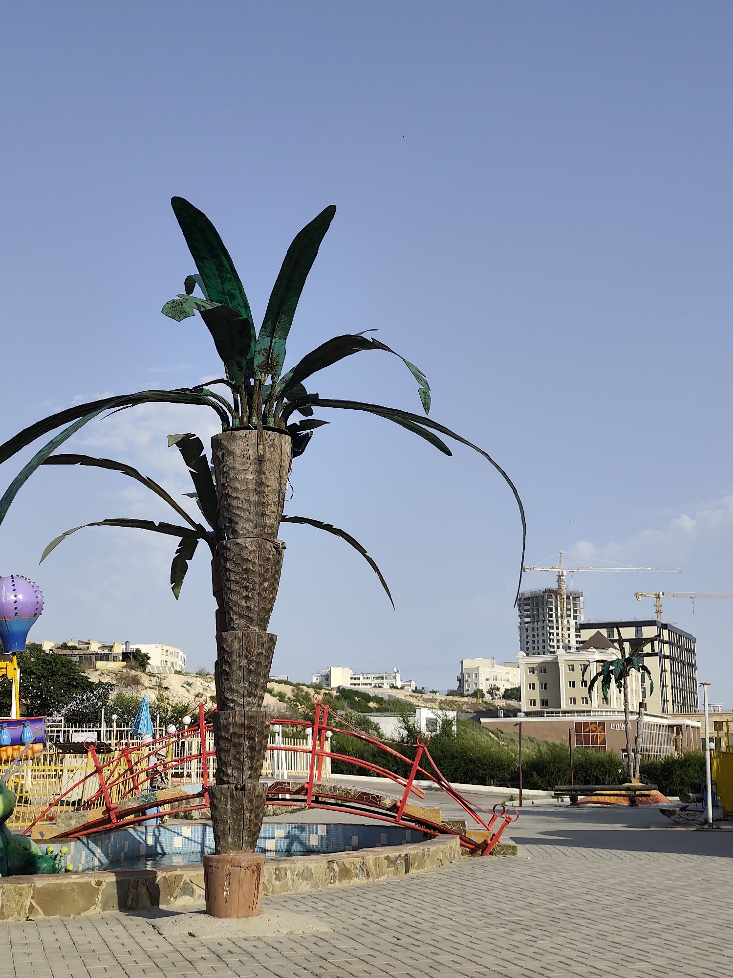The image depicts an outdoor scene on a clear day, likely in a public space. In the foreground, a large, artificial palm tree sculpture made of wood and metal stands prominently. The sculpture has a textured, cylindrical trunk and metallic green leaves. The base of the sculpture is set in a small, decorative fountain with tiled edges and stone accents.
Behind the sculpture, a red and yellow bridge arcs over the fountain. To the left, part of a colorful playground attraction with a purple balloon-like element is visible. In the mid-ground, there are more playground elements, including a white fence and a red railing. Beyond this, a landscaped area with green bushes and trees slopes upwards.
In the background, the urban landscape of Aktau, Kazakhstan, is evident. Several buildings are under construction, marked by large cranes. One building is a multi-story structure with a grey, unfinished exterior, while another is a completed, modern building with dark windows and an angular design. Other residential or administrative buildings are visible on the hillside. The sky is a clear, pale blue with a few faint clouds. The ground in the foreground is paved with light-colored rectangular blocks.