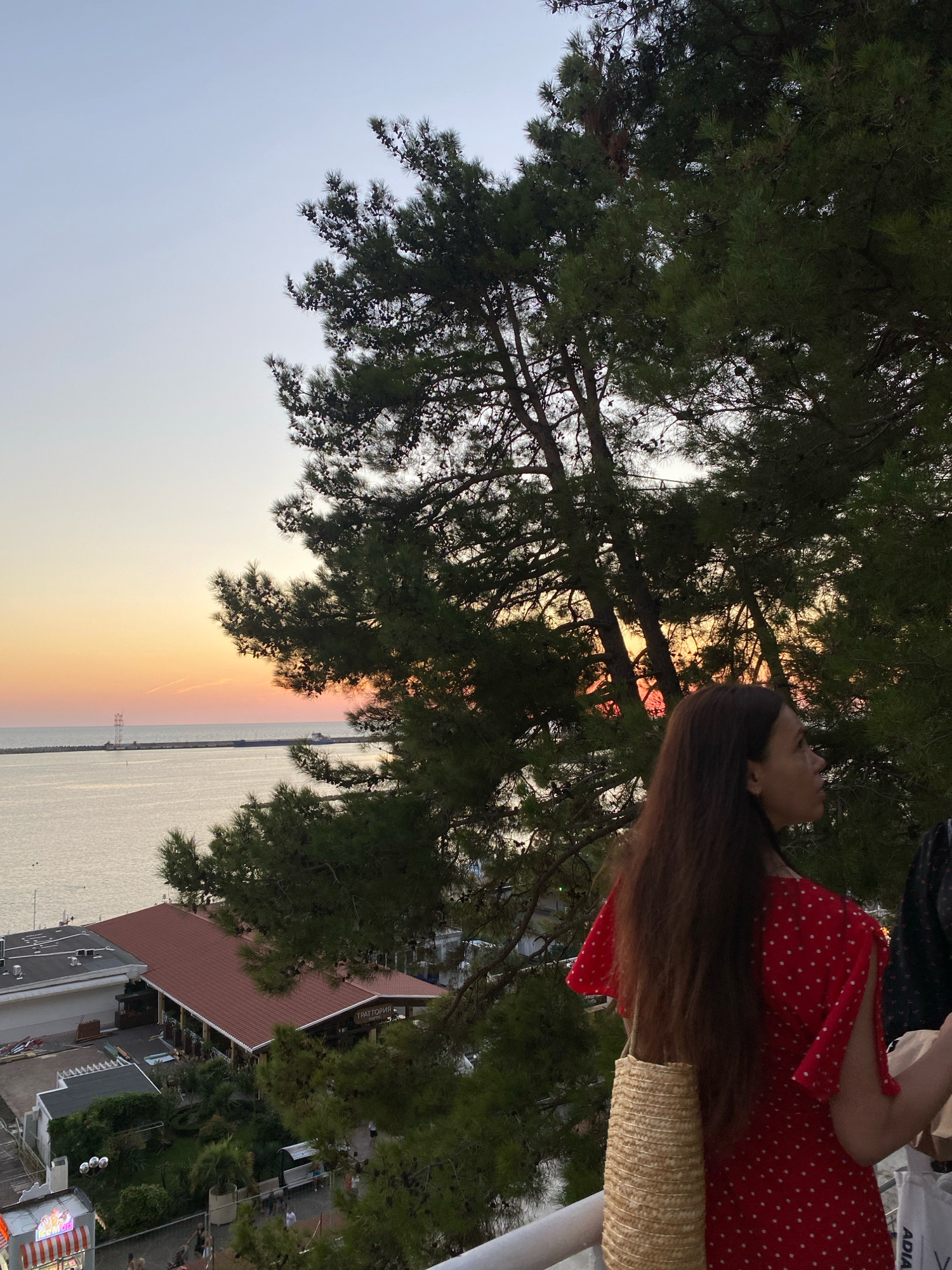An outdoor medium shot captures a young woman with long, dark brown hair standing in front of a backdrop of pine trees and a sunset over the Black Sea. She is wearing a red dress with white polka dots and a straw bag slung over her shoulder. To her right, another person is partially visible. The scene is set in Sochi, Russia, with buildings and a pier visible in the distance along the coastline. The sky displays a gradient of colors, from light blue at the top to orange and pink near the horizon, indicative of a sunset.